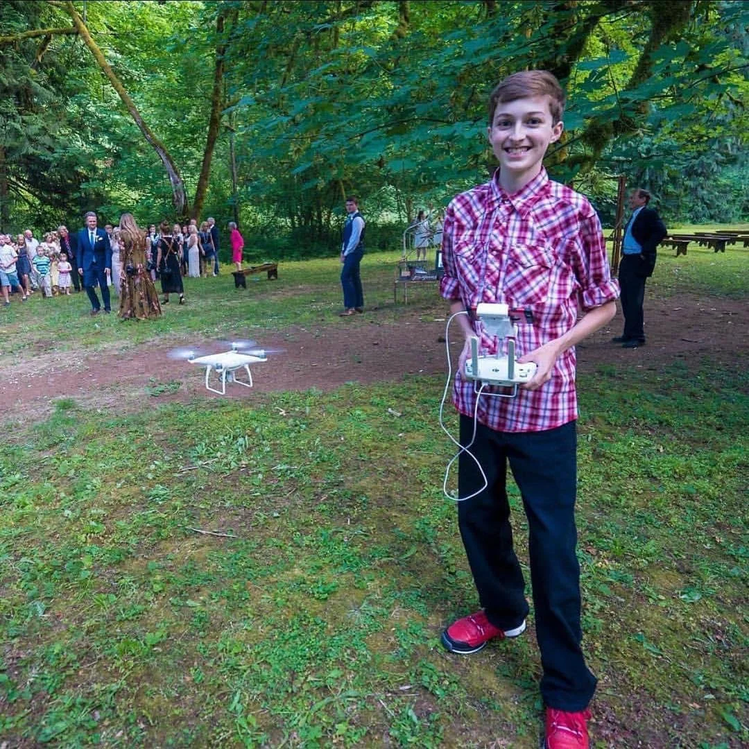 A smiling young boy in a red checkered shirt and black pants is standing outdoors in a forested area, holding a drone controller. Behind him, a drone is flying close to the ground. In the background, a group of people dressed in formal attire are gathered, possibly at a wedding or celebration, with trees and greenery surrounding them.