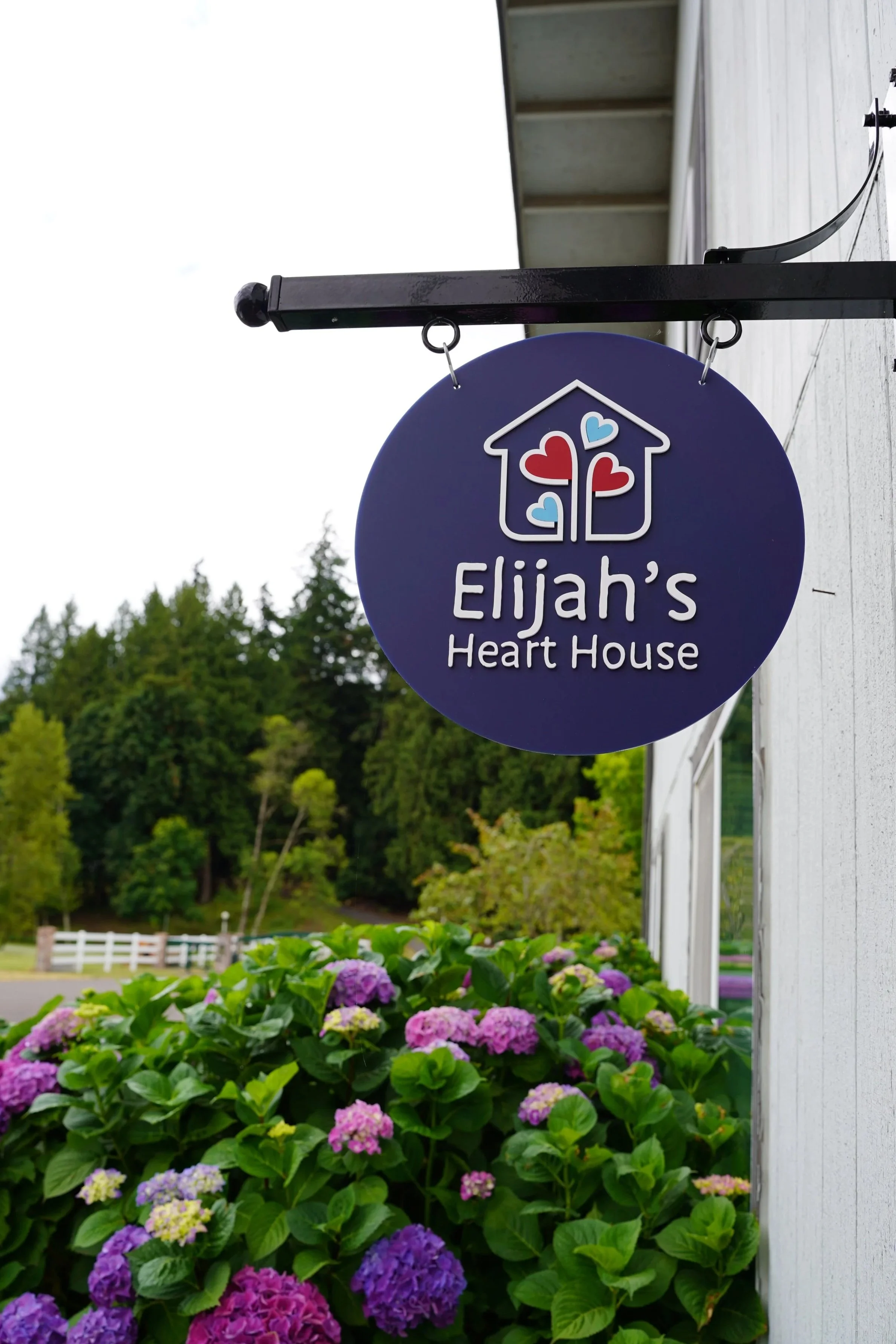 Sign for Elijah's Heart House hanging on black metal bracket outside a white building, with blooming purple and pink hydrangea flowers in the foreground and green trees in the background.