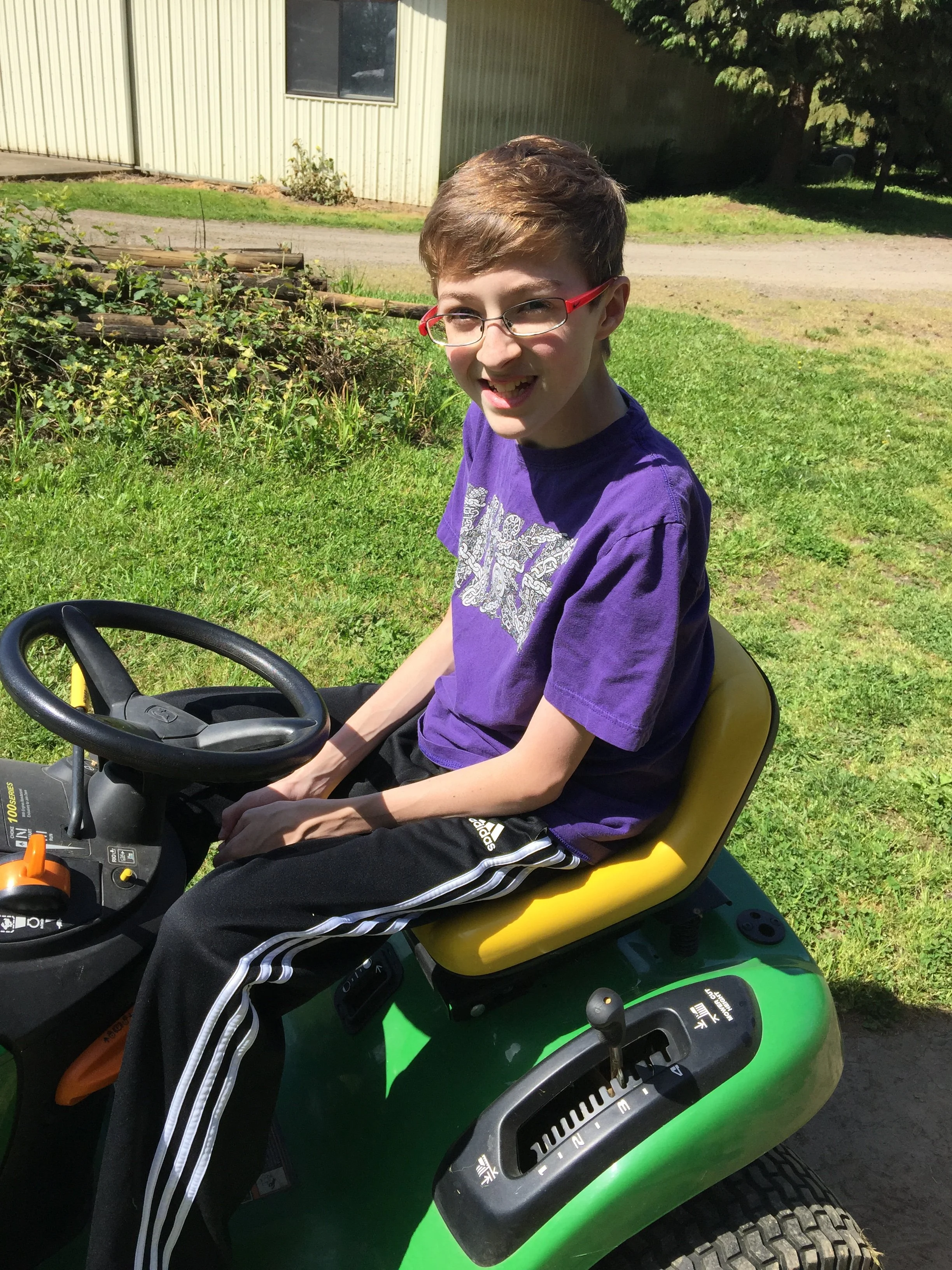 A young boy with glasses smiling while sitting on a green lawn tractor in a grassy area outdoors, with a building and trees in the background.