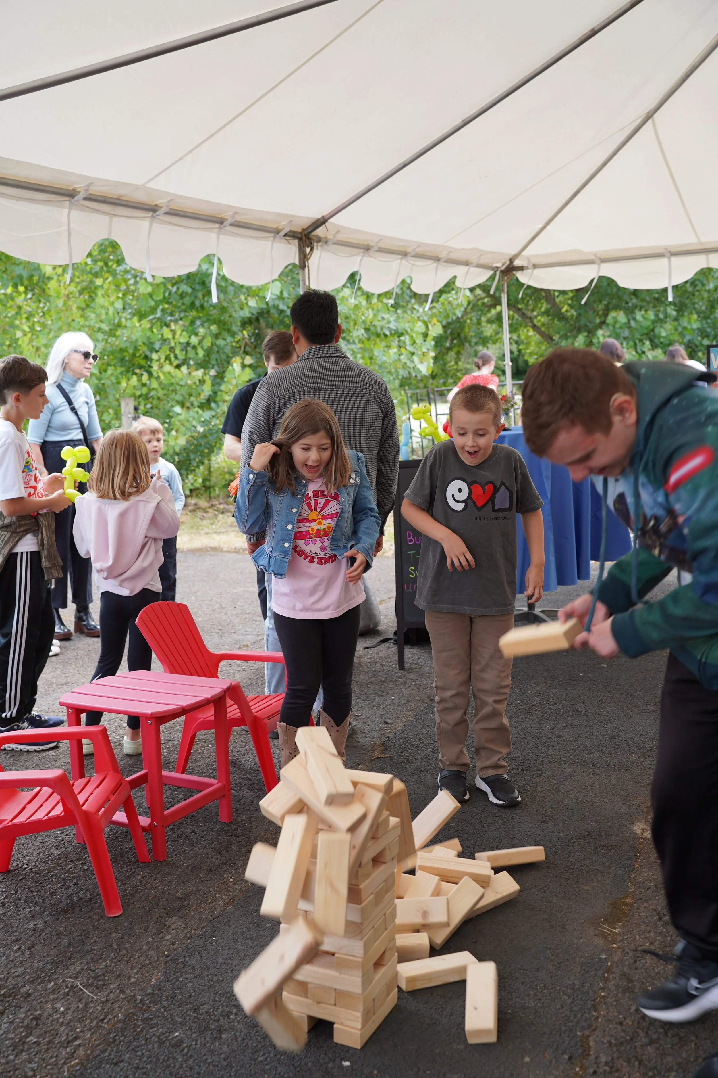 Children playing Jenga at an outdoor event under a large white canopy with green trees in the background.
