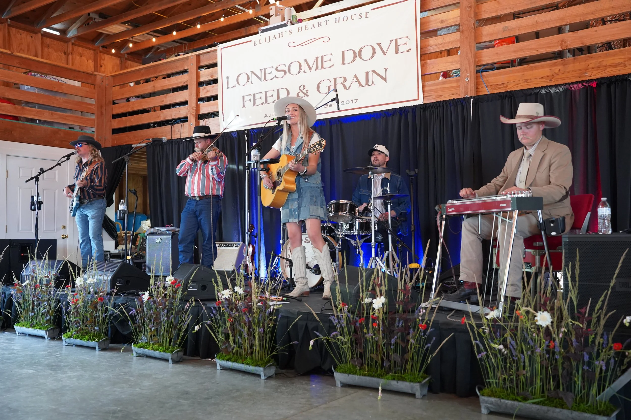 A live band performing on stage at an indoor event with a wooden roof and decor. The band includes a woman singer with a guitar, a man playing violin, a drummer, and a keyboard player all dressed in country or western attire. The stage is decorated with flowers in pots along the front.