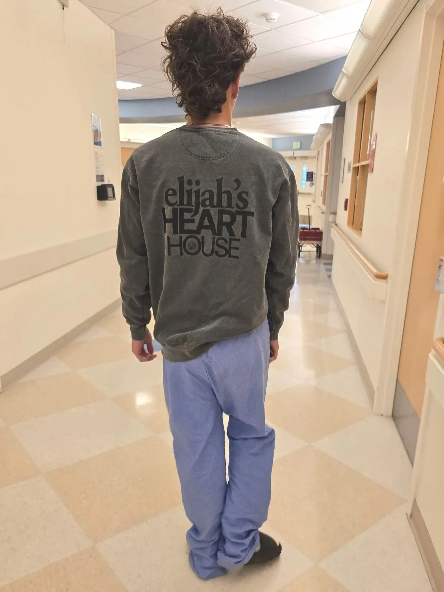 Person walking through a hospital corridor, wearing a gray sweatshirt with the words 'elijah's HEART HOUSE' on the back, light blue hospital pants, and black shoes.