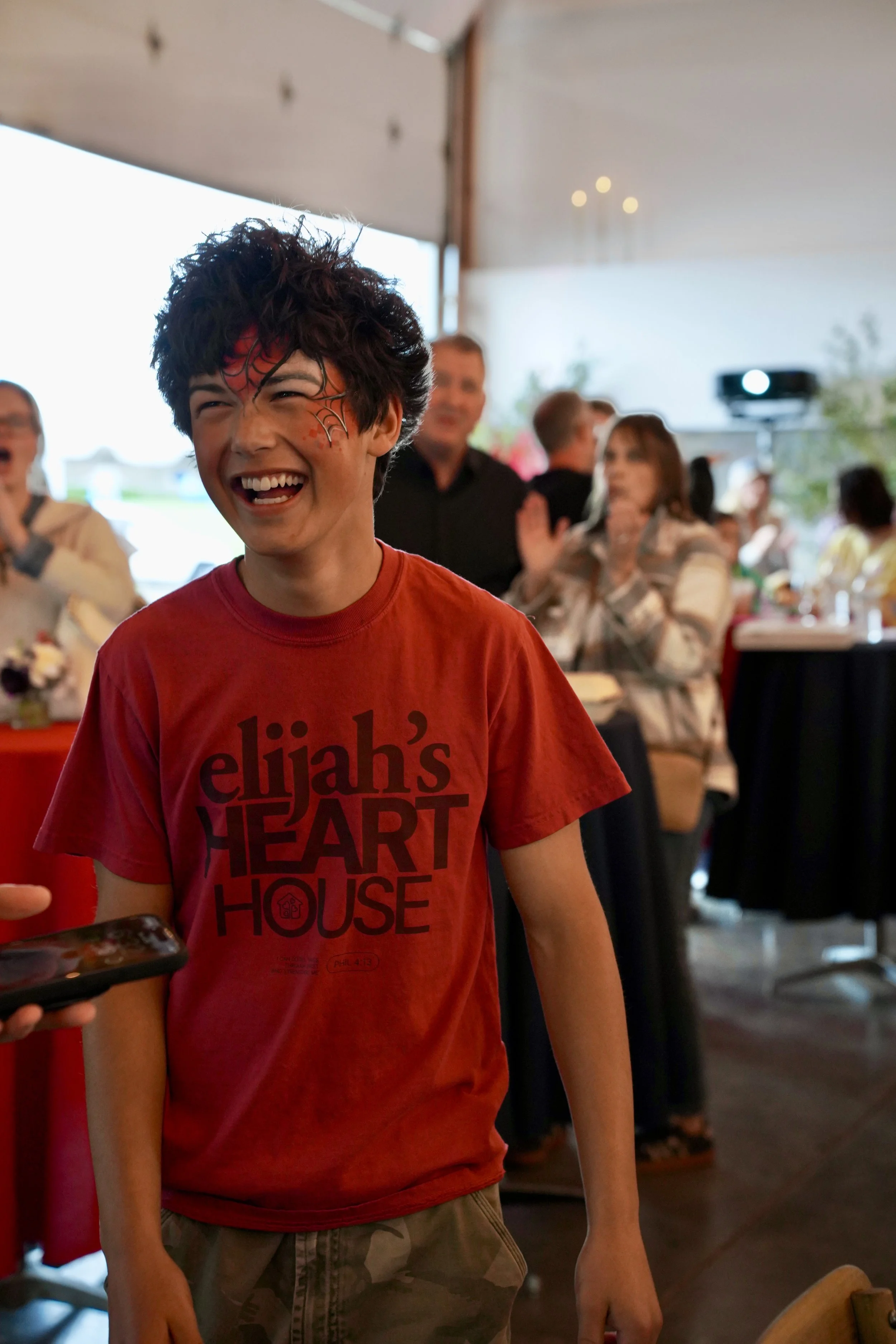Boy in a red Elijah's Heart House shirt with painted spiderman face smiling.