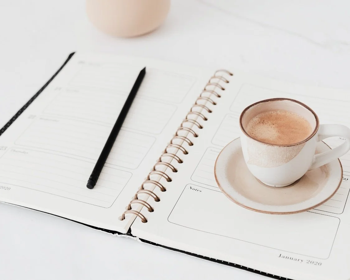 Open planner with blank pages, a black pen, a ceramic coffee cup with frothy coffee on a matching saucer, on a white surface.