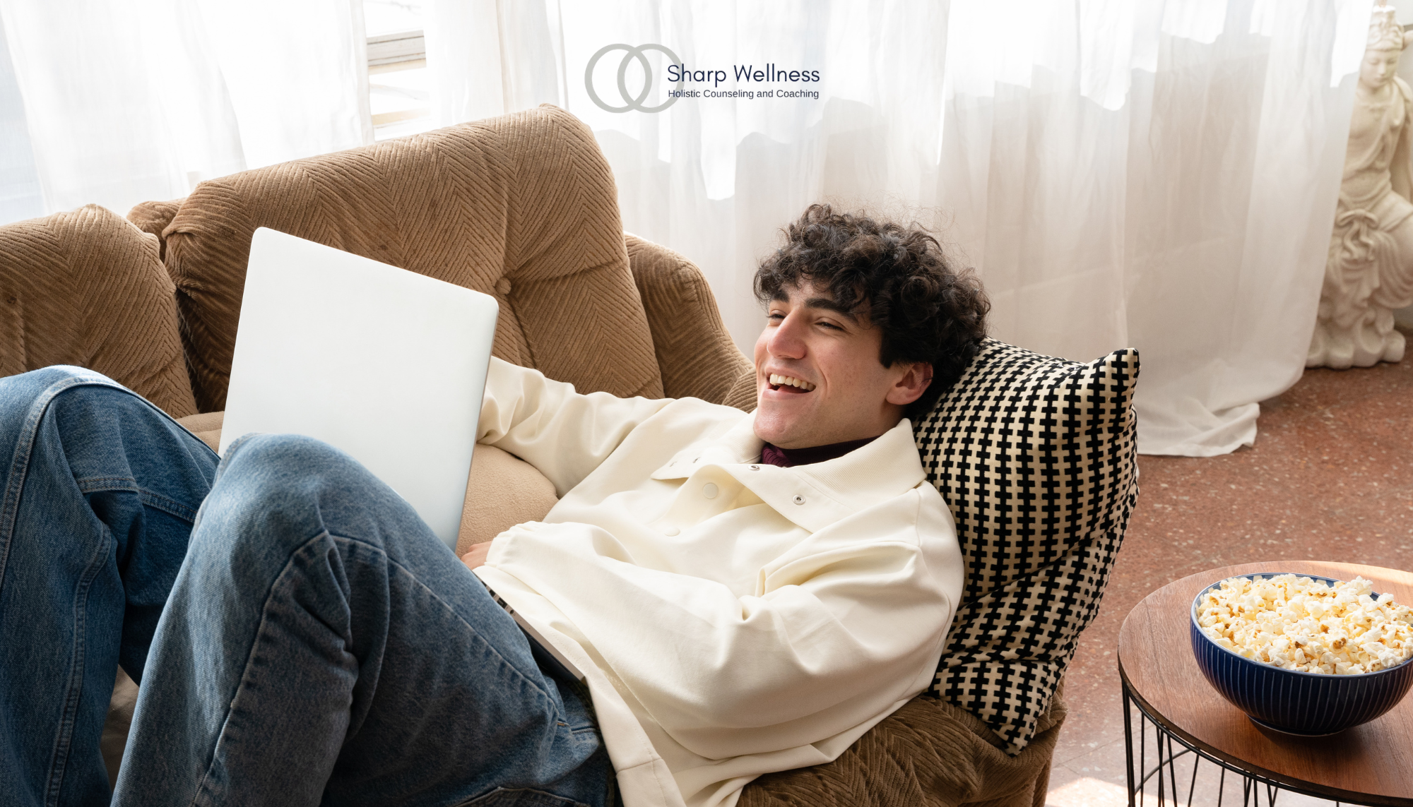 Young man relaxing on a couch with a laptop and popcorn, representing watching horror movies for entertainment and stress relief.