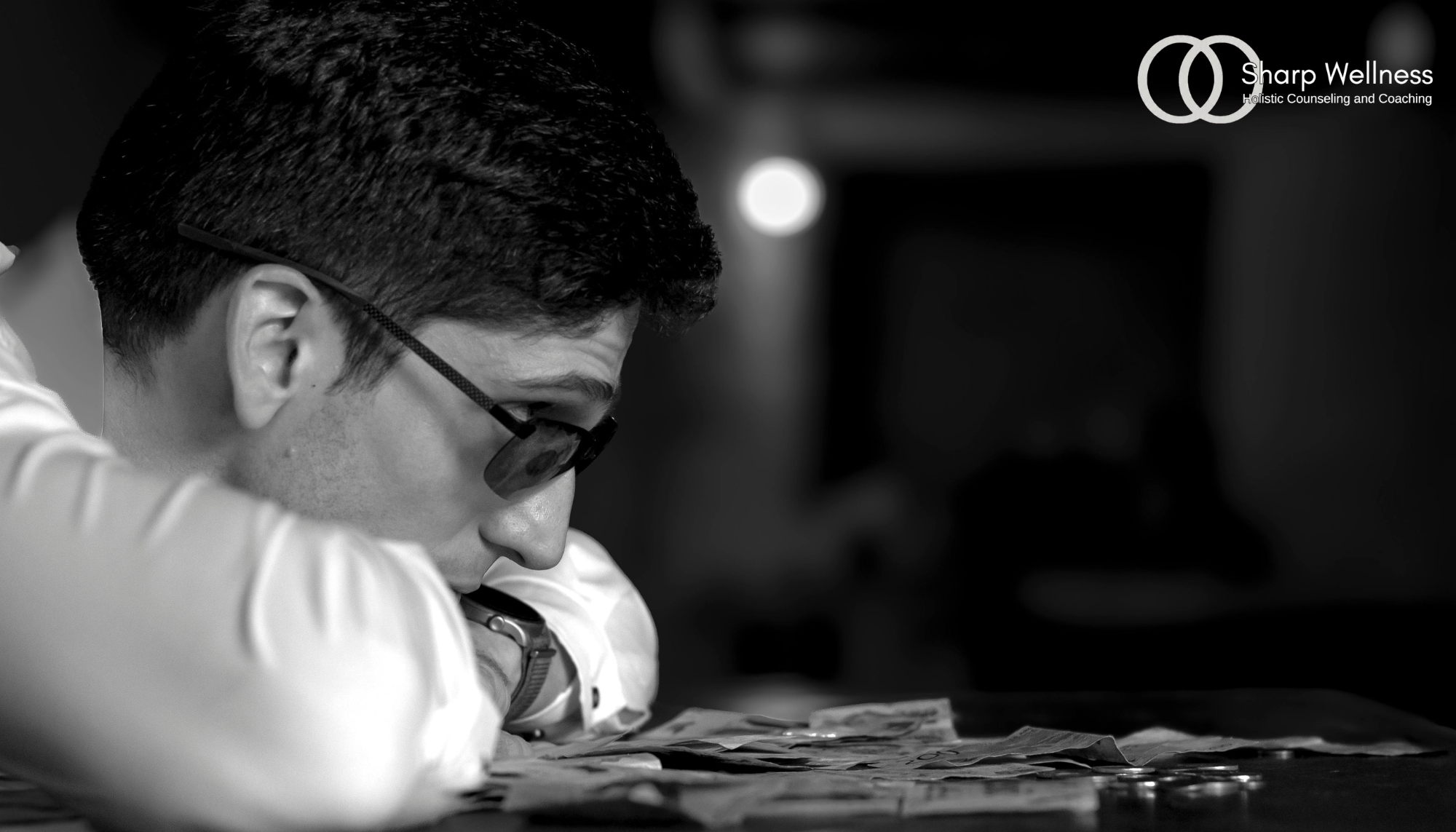 Man in sunglasses leaning over a table with scattered money, illustrating stress, fixation, or intrusive thoughts.