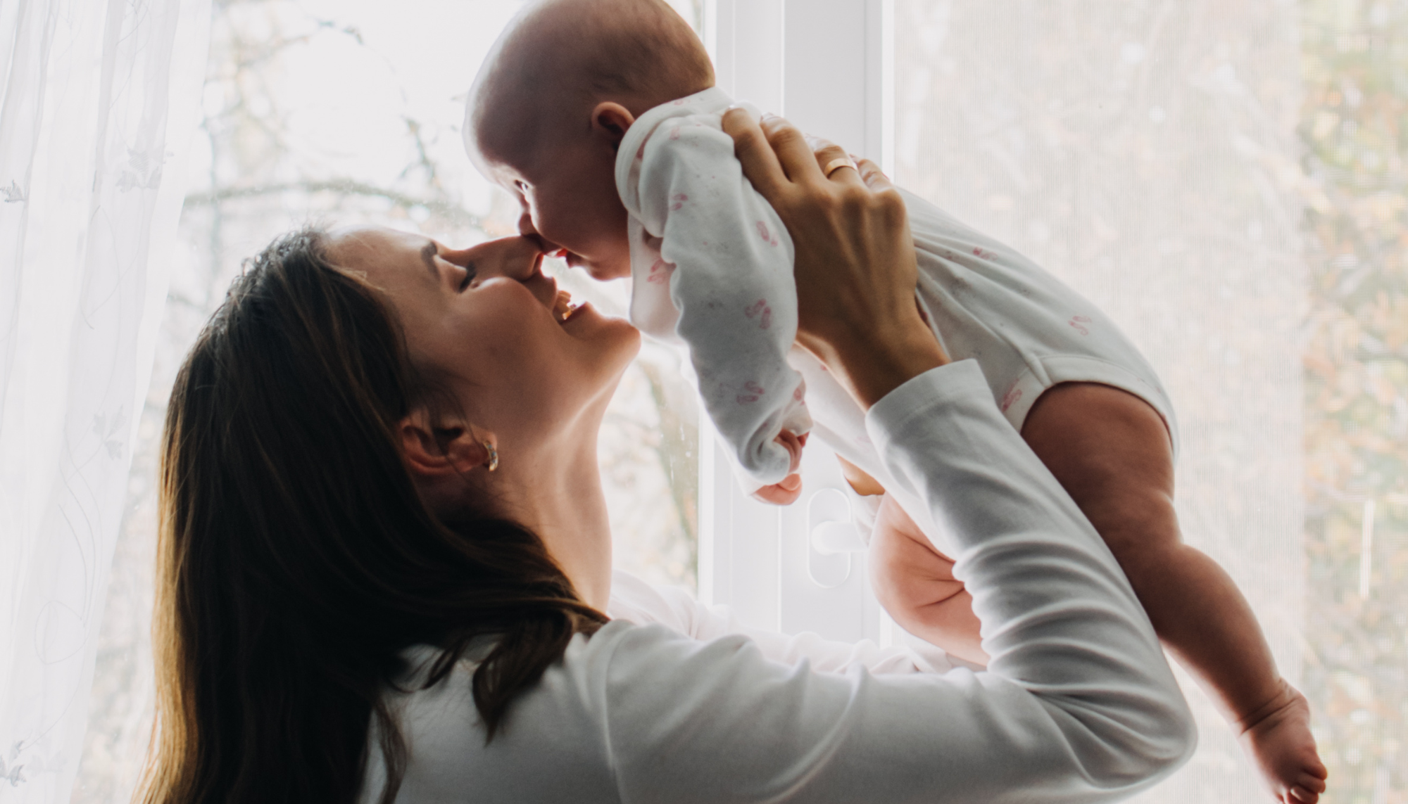 A mother smiles while holding her baby near a bright window, representing postpartum support, maternal mental health, and the emotional connection of early motherhood.
