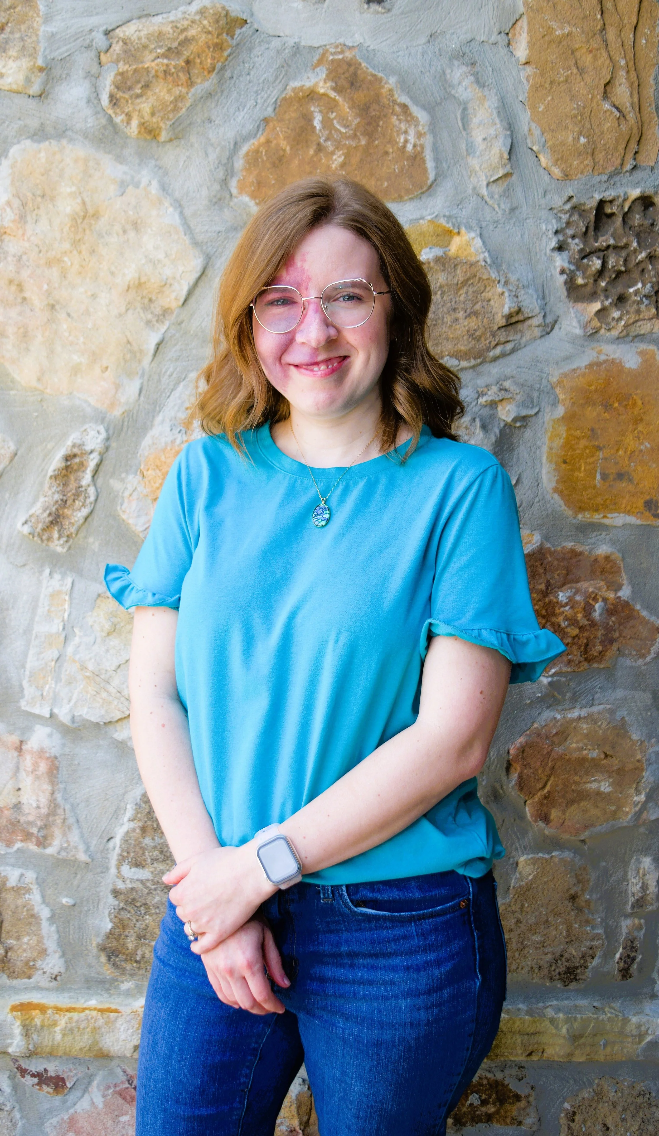 Rachel McCullough, PhD, LPC, PMH-C, Senior therapist at Sharp Wellness Counseling Vestavia Hills, Alabama. Pictured is a young woman with short red hair, glasses, and a blue shirt with ruffled sleeves, smiling outdoors with stone in the background.