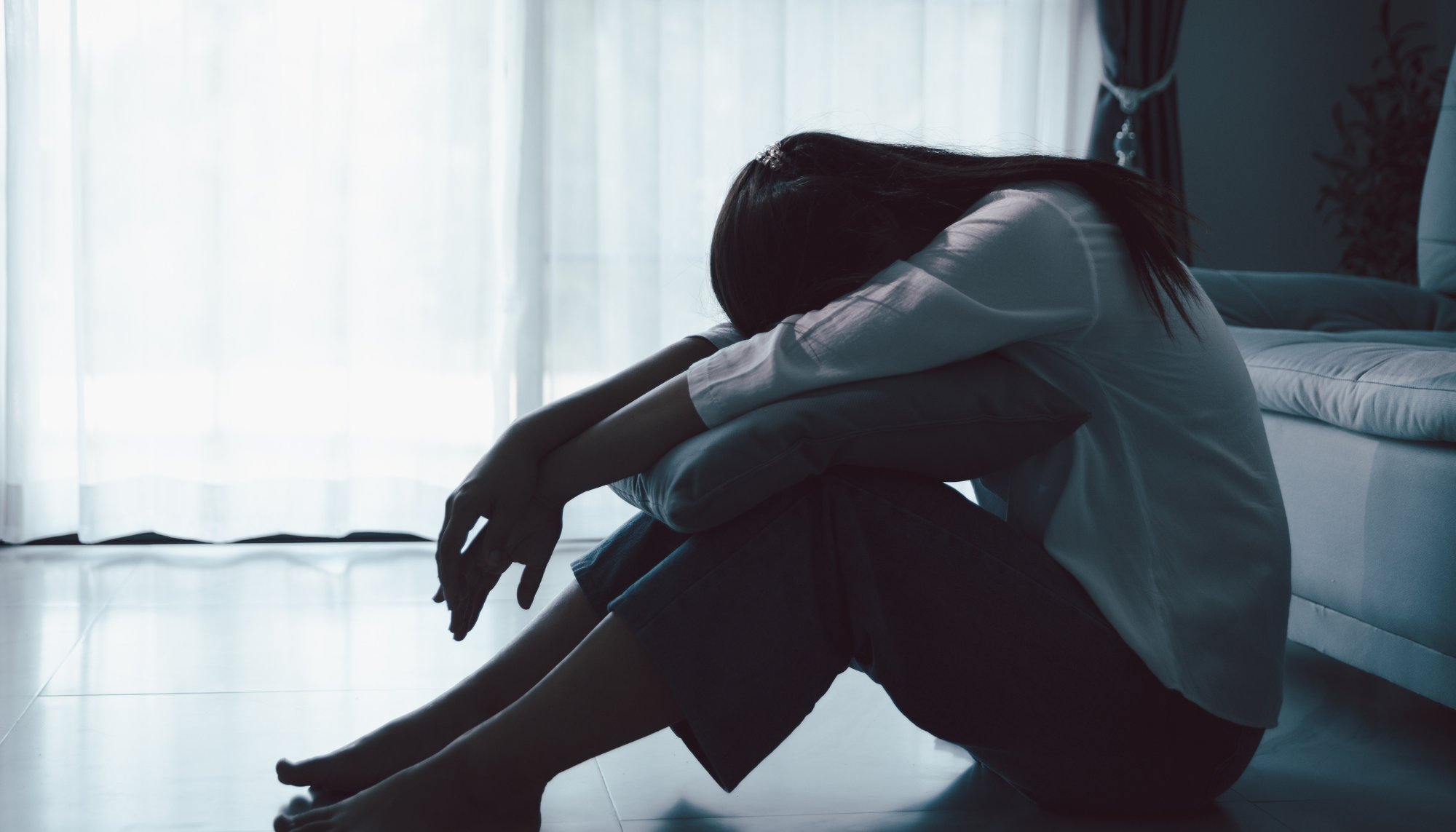 Woman sitting on the floor with her head down near a couch, representing depression, sadness, isolation, and feeling overwhelmed.