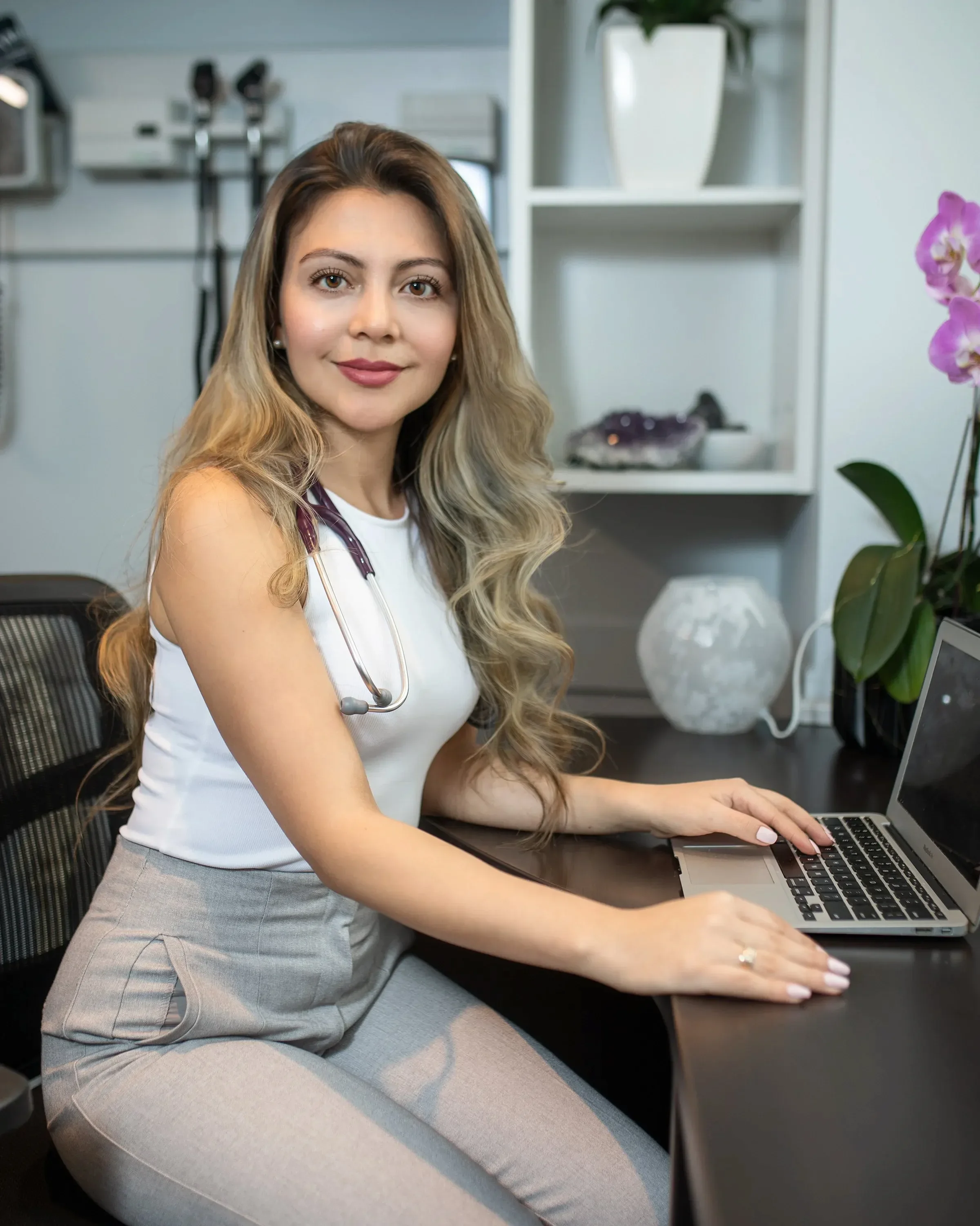 A young woman with long wavy blonde hair wearing a stethoscope around her neck, white top, and gray pants, sitting at a desk and working on a laptop in a modern, well-lit office or medical setting.