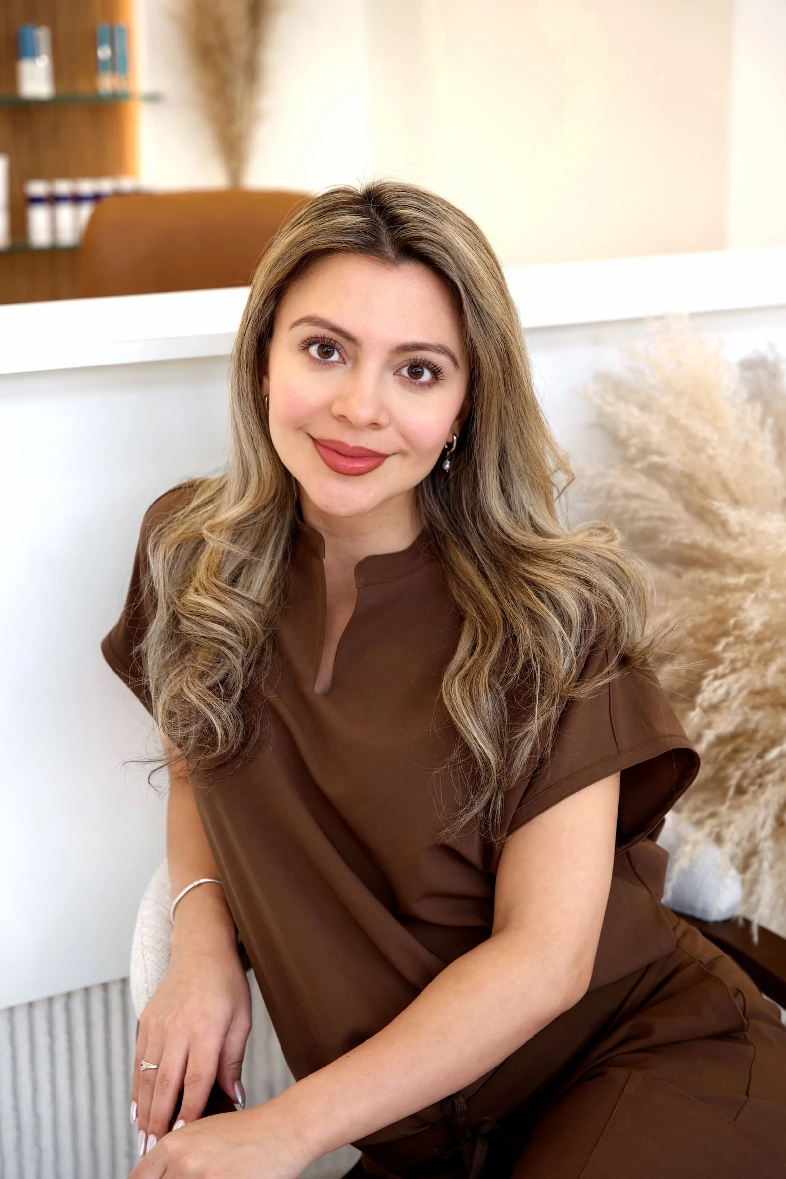 A young woman with long, wavy light brown hair and brown eyes, wearing a brown top, sitting indoors in front of a beige wall and white shelf, with a relaxed and friendly expression.