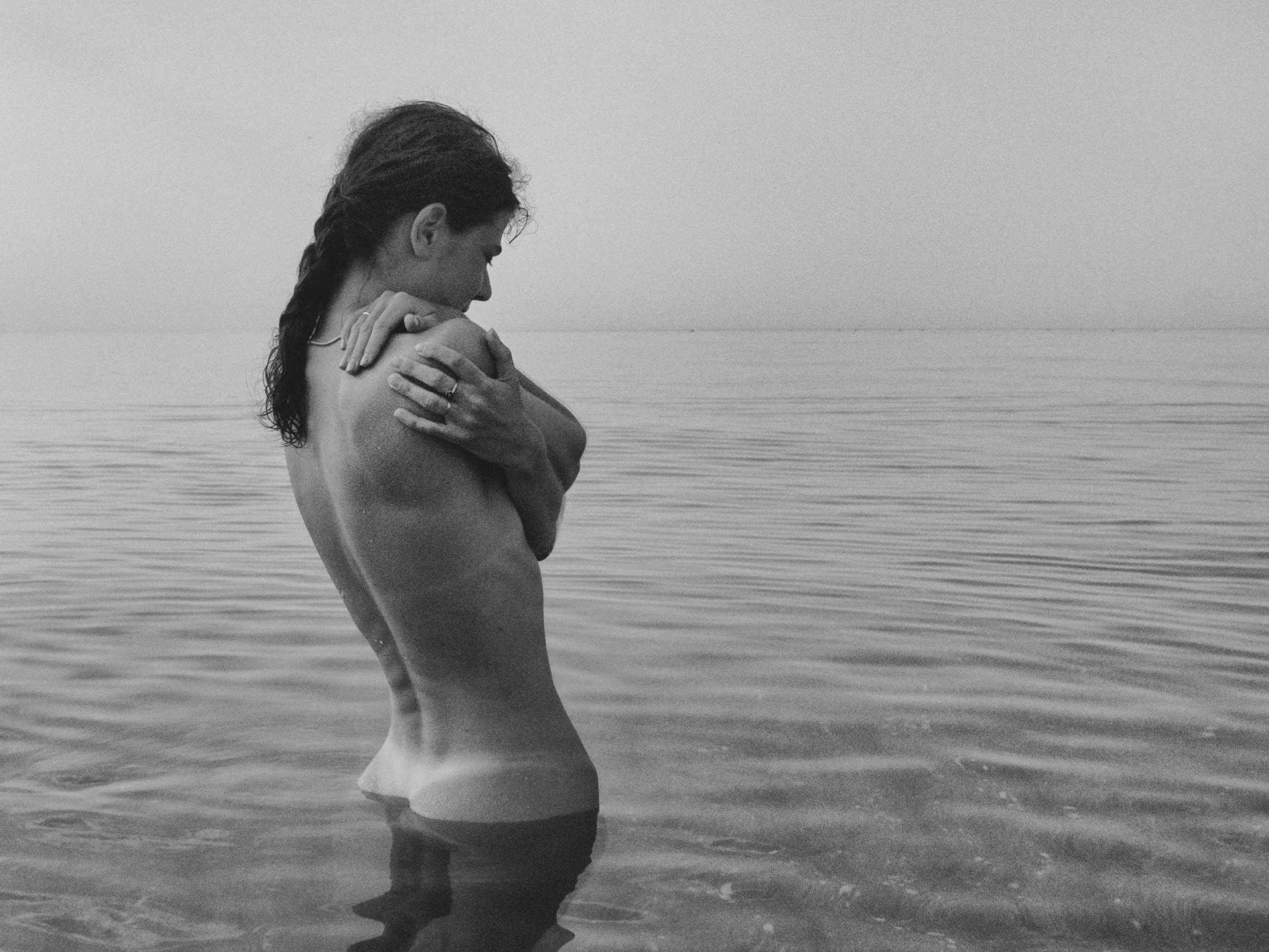 A person with long hair tied back, standing in shallow water at the beach, holding their shoulder with one hand, in a contemplative pose, with the horizon and calm water in the background.