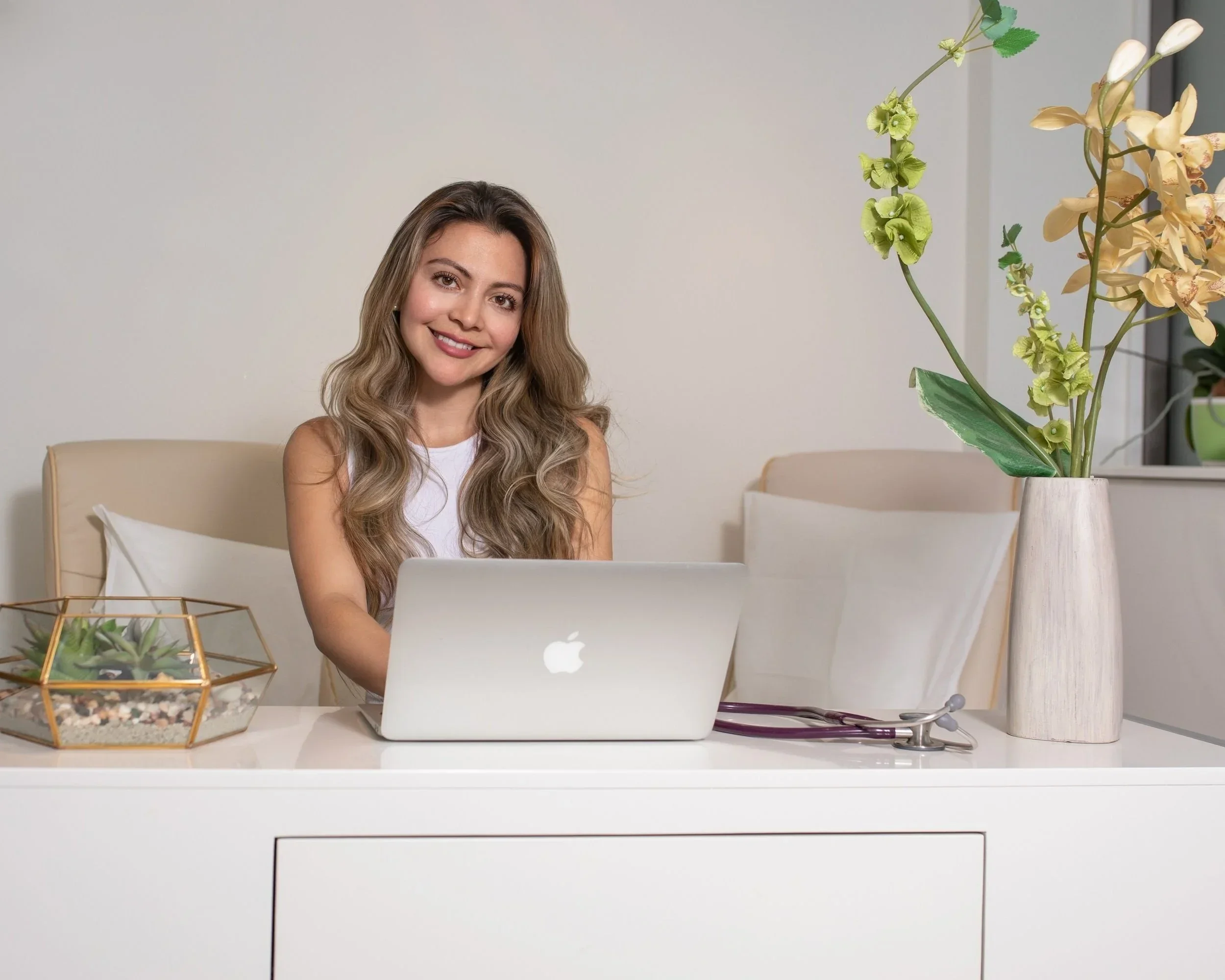A young woman with wavy blonde hair and a white sleeveless top sitting at a white desk with a silver MacBook in front of her. There is a small glass terrarium with plants to her left, and a large bouquet of white and green flowers in a white vase to her right. Behind her is a beige chair with a white pillow, and the background features a light-colored wall and some shelves.