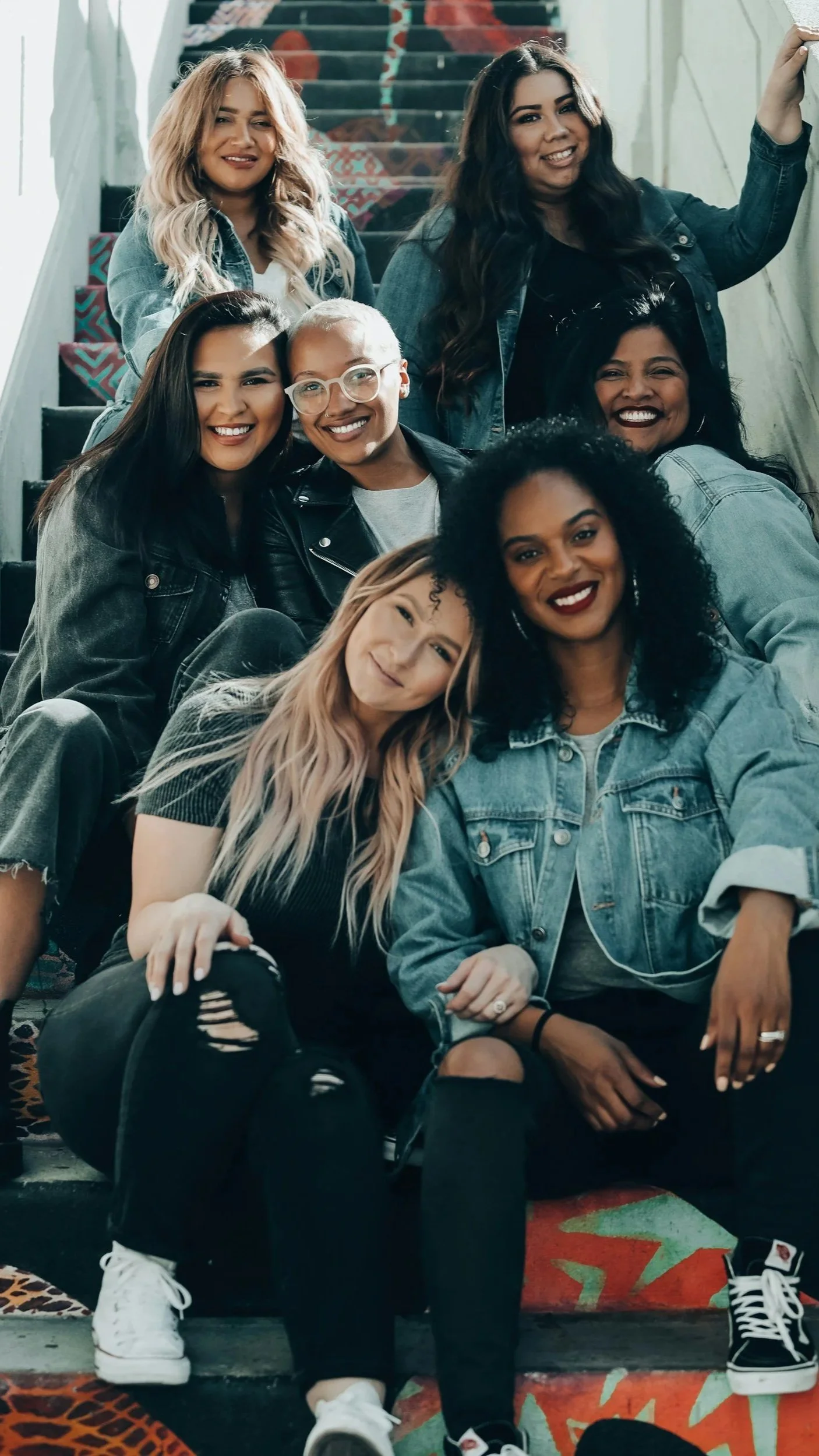 A group of seven women of diverse ethnicities smiling and sitting on outdoor stairs, with some leaning on each other, wearing casual clothes like denim jackets and black pants.