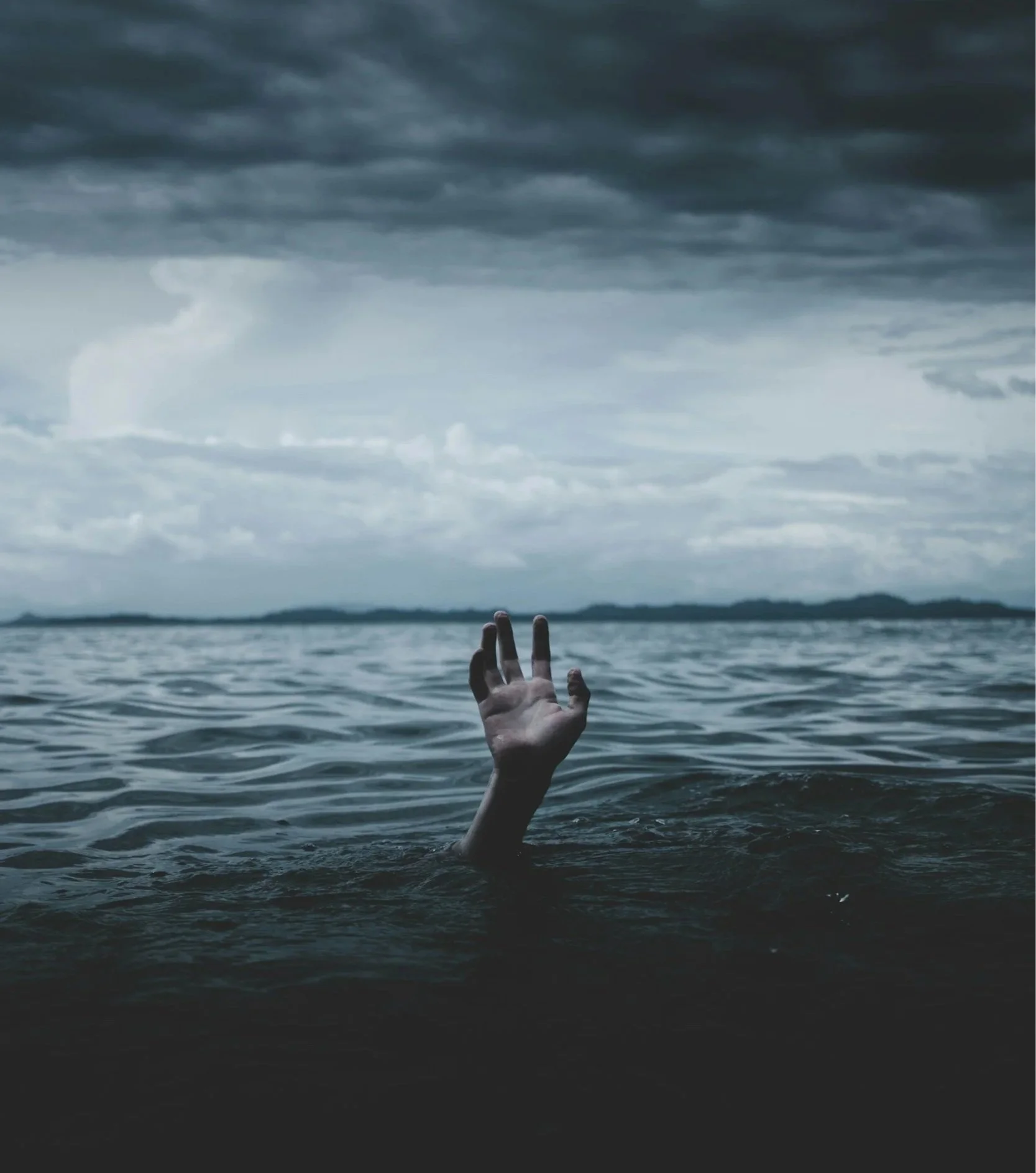 A person's hand is reaching up through the surface of a body of dark water. The sky is overcast with dark clouds, and the horizon is visible in the distance.