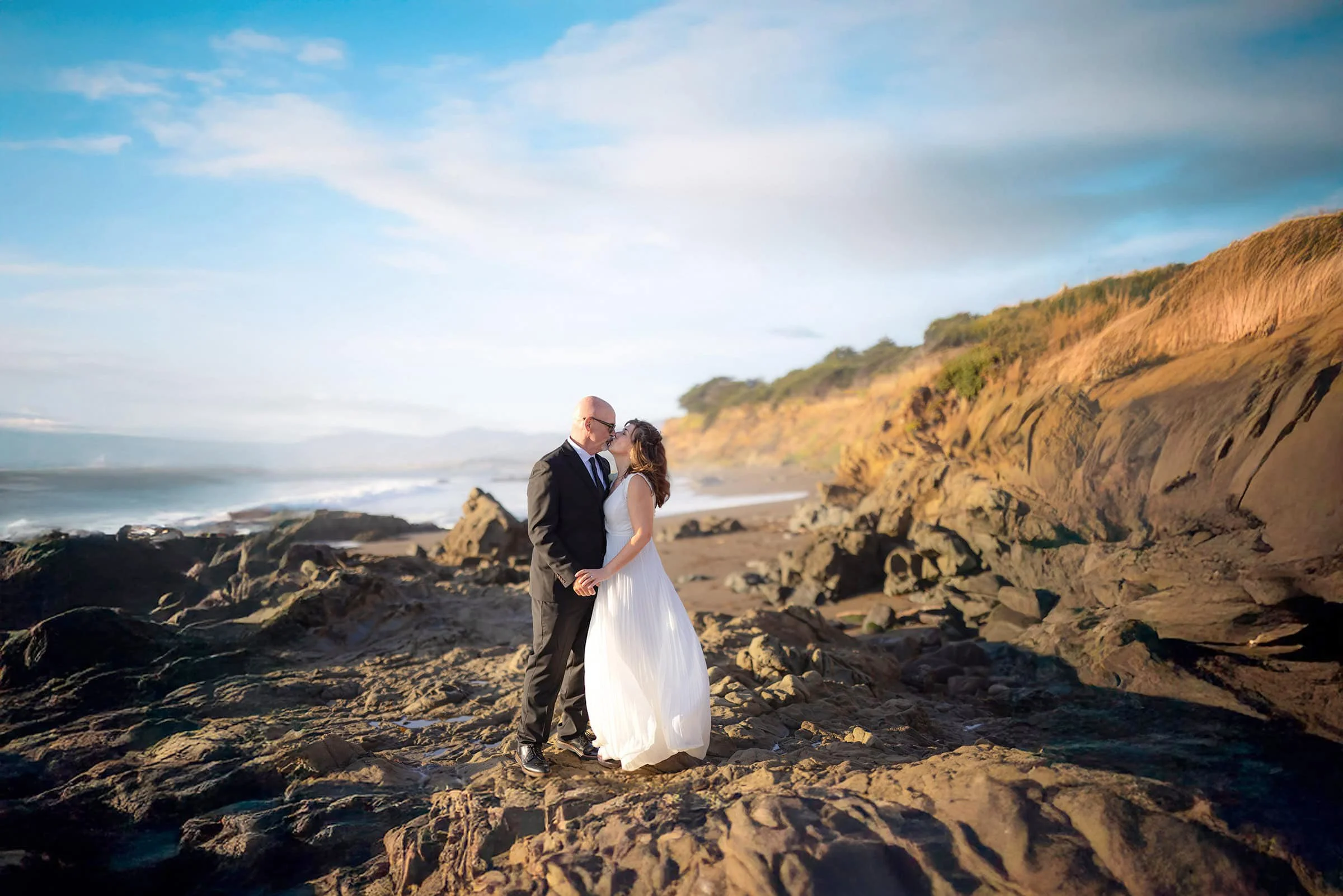 Bride and groom embracing on jagged coastal cliffs in Cambria, California, with blue skies and waves crashing below