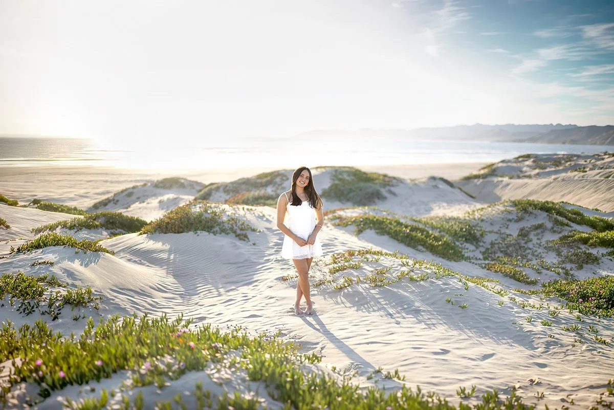 Cal Poly senior standing among the Pismo Beach dunes with sunlight shining through the ice plant