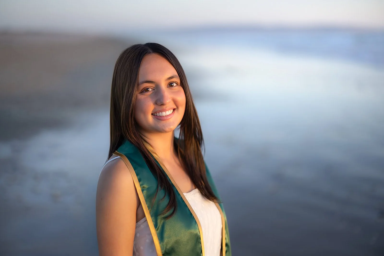 A young woman with long brown hair smiling at the camera, standing near a body of water during sunset, wearing a graduation stole.