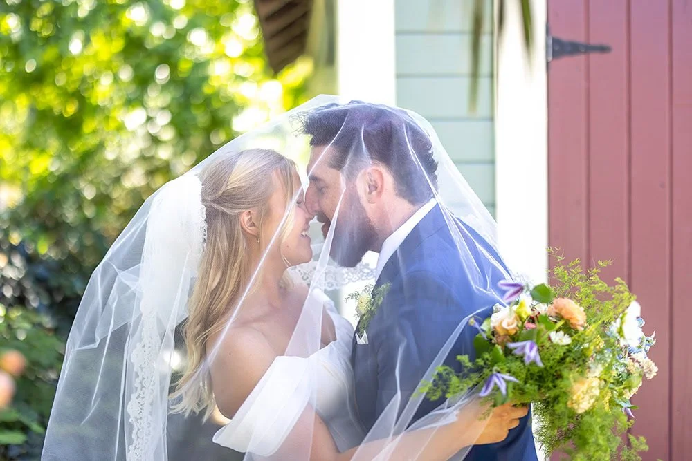 Bride and groom touching noses beneath the bride’s veil as she holds her bouquet, with sunlight beaming through the trees behind them at Heritage House in Arroyo Grande, California