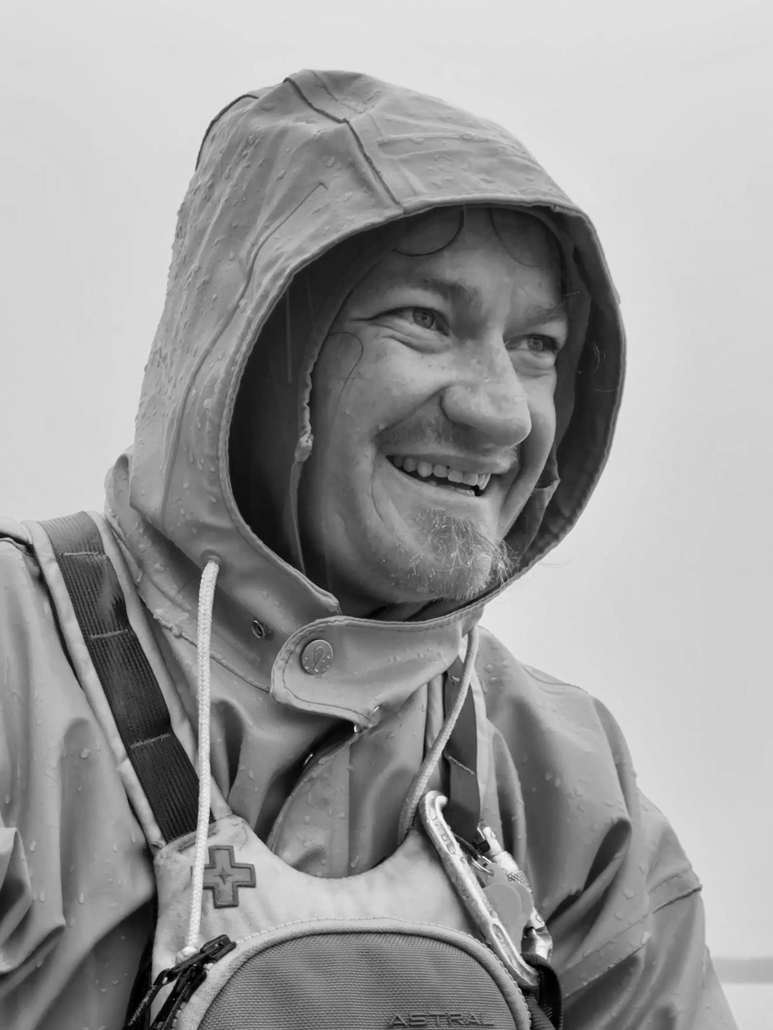 A man wearing sunglasses and a visor hat, smiling in a mountainous outdoor setting in black and white.