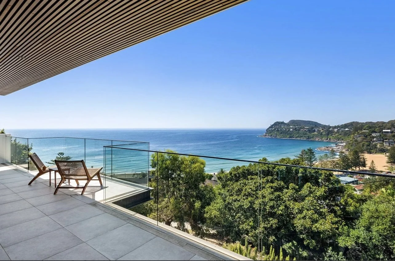 Balcony with two chairs overlooking a beach, ocean, and greenery under a clear blue sky.