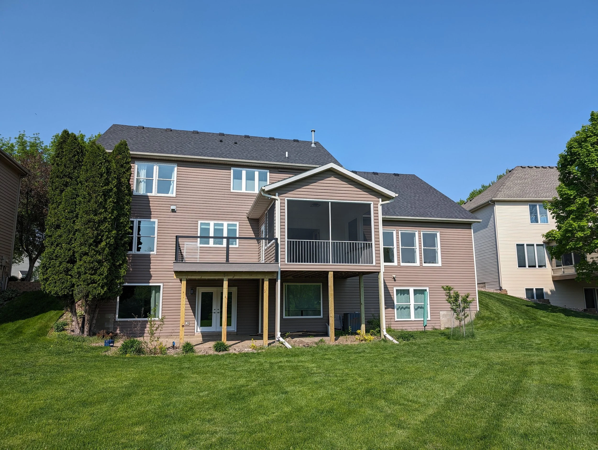 Back view of a multi-story house with a covered patio and a balcony, surrounded by green grass and trees, under a clear blue sky.