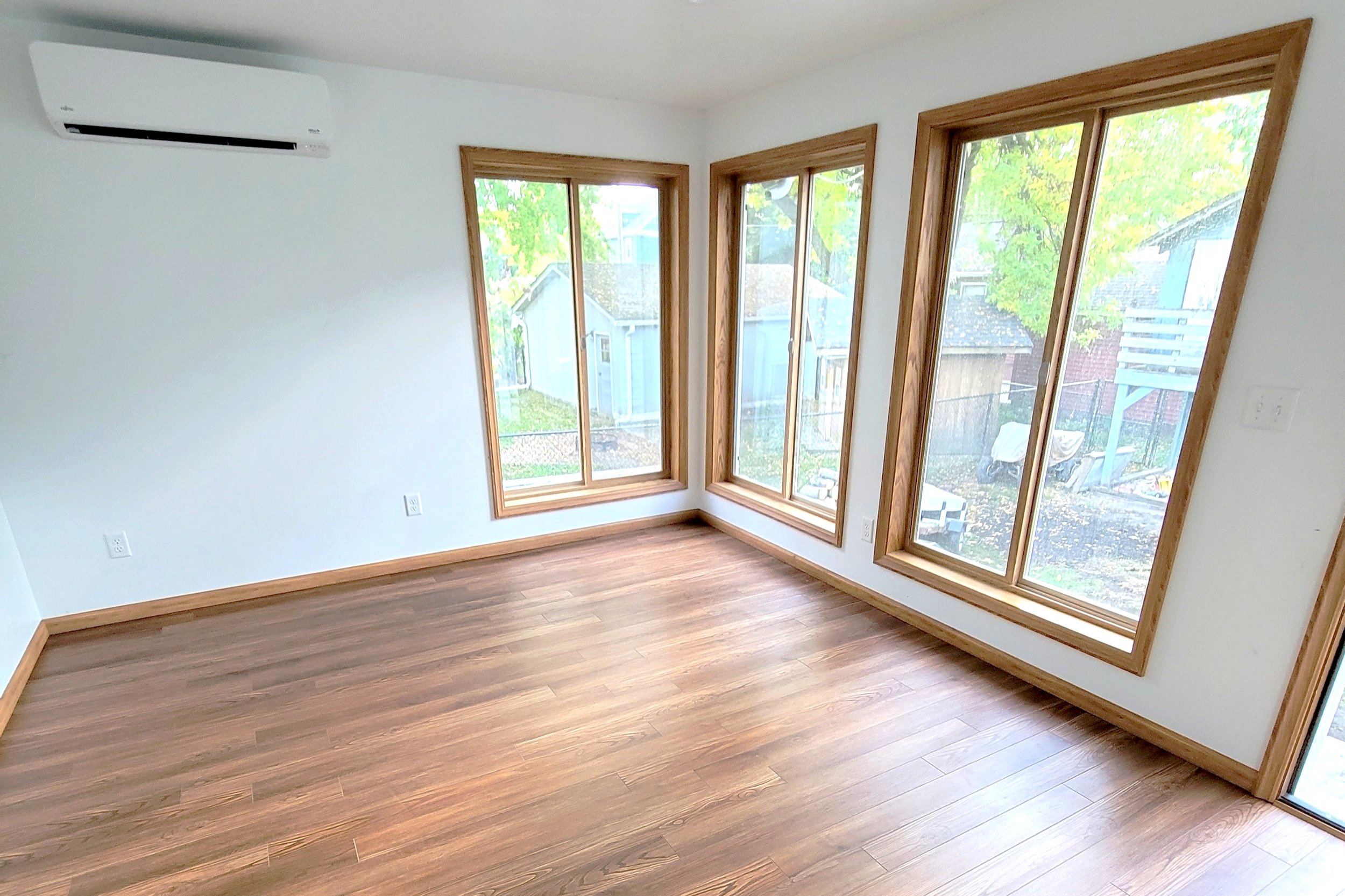 Empty room with hardwood floors, white walls, three large windows with wooden frames, and an air conditioning unit on the wall.