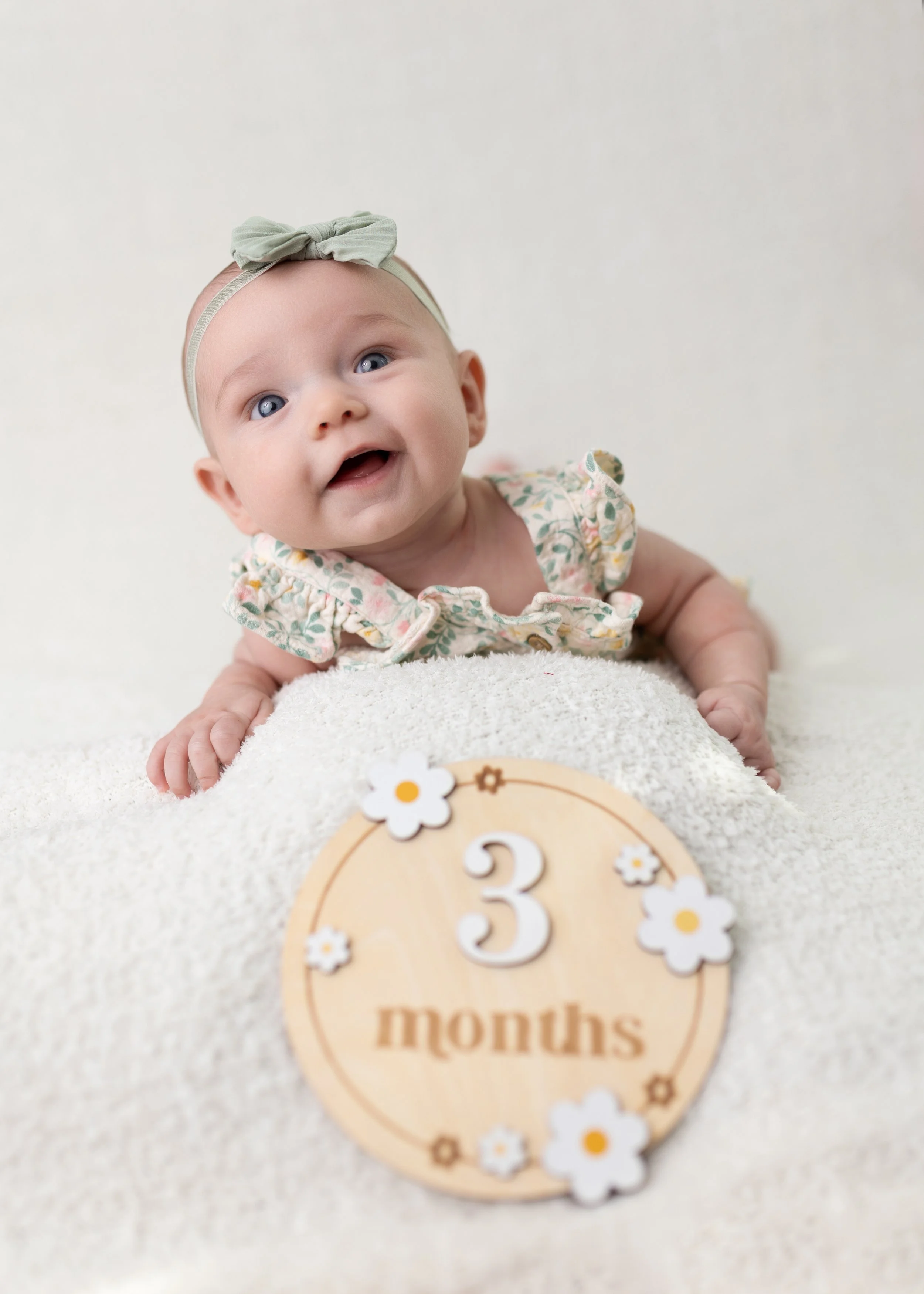 A smiling baby girl with a bow headband and floral dress lying on her stomach on a soft surface, next to a wooden decoration with the number three and the word 'months' for a milestone photo.