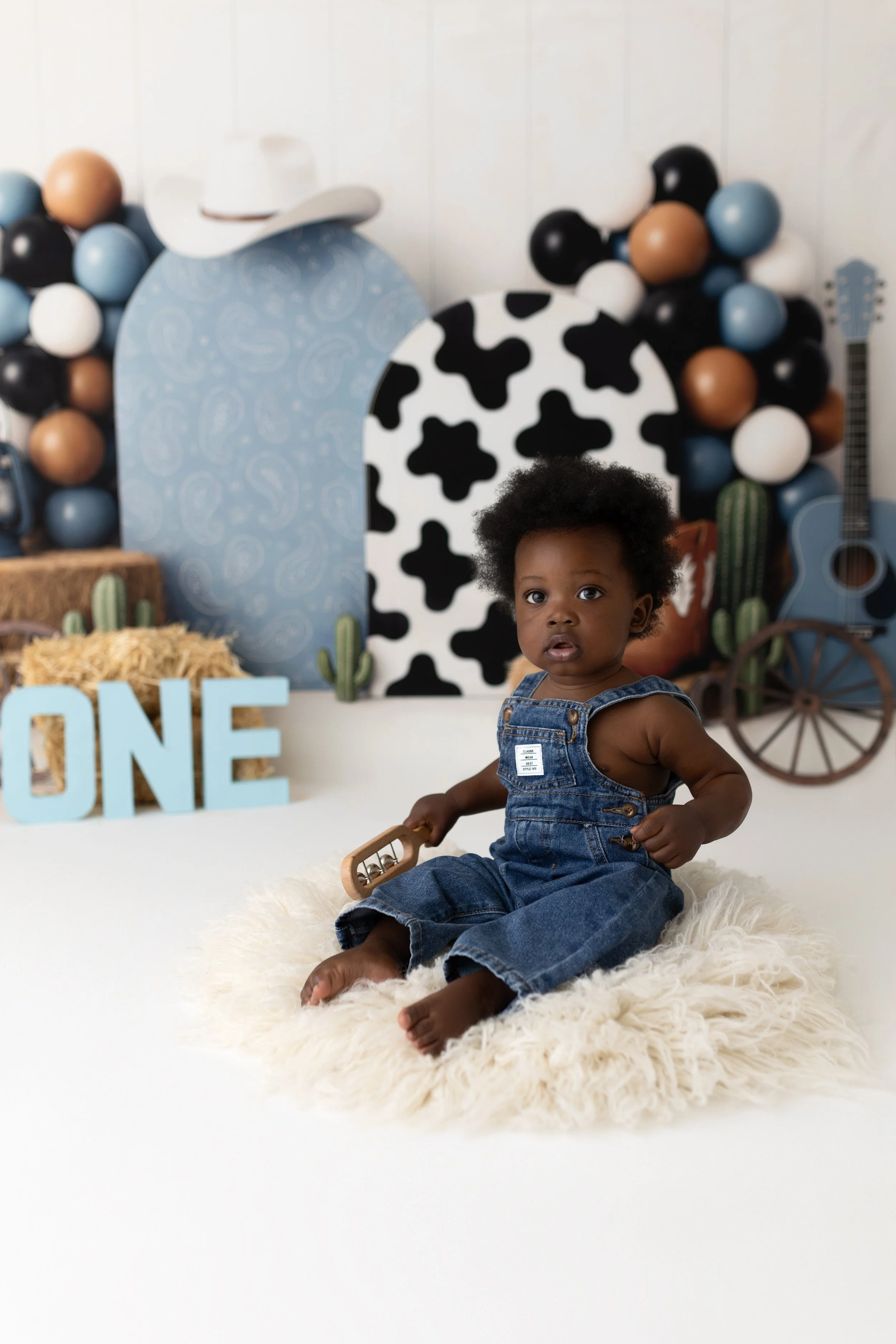 A one-year-old child sitting on a fluffy rug, holding a wooden toy, surrounded by a country-themed backdrop with balloons, cacti, and a sign that reads 'ONE'.