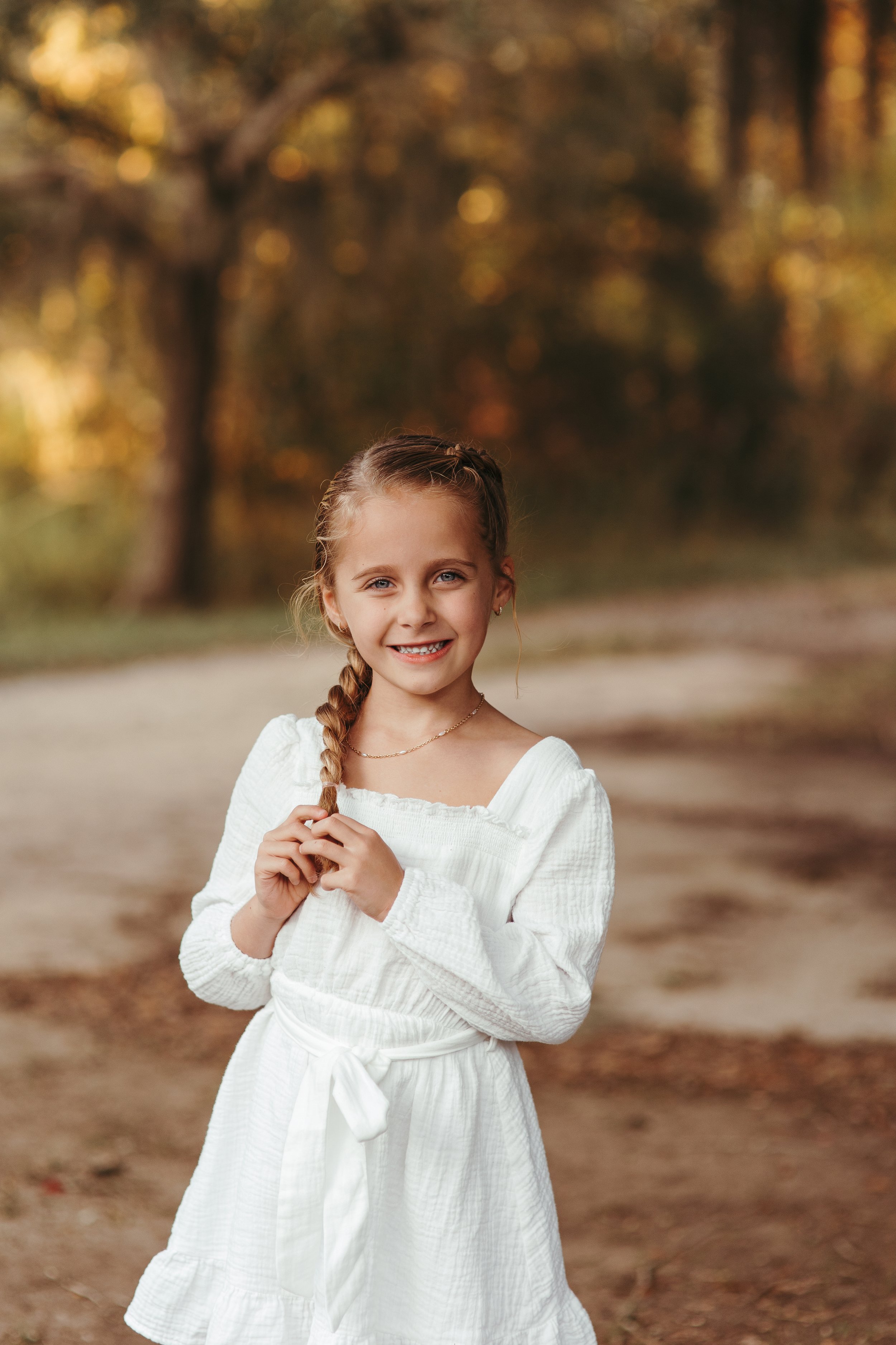 A young girl with braided hair wearing a white dress, standing outdoors in a wooded area during autumn, smiling at the camera.