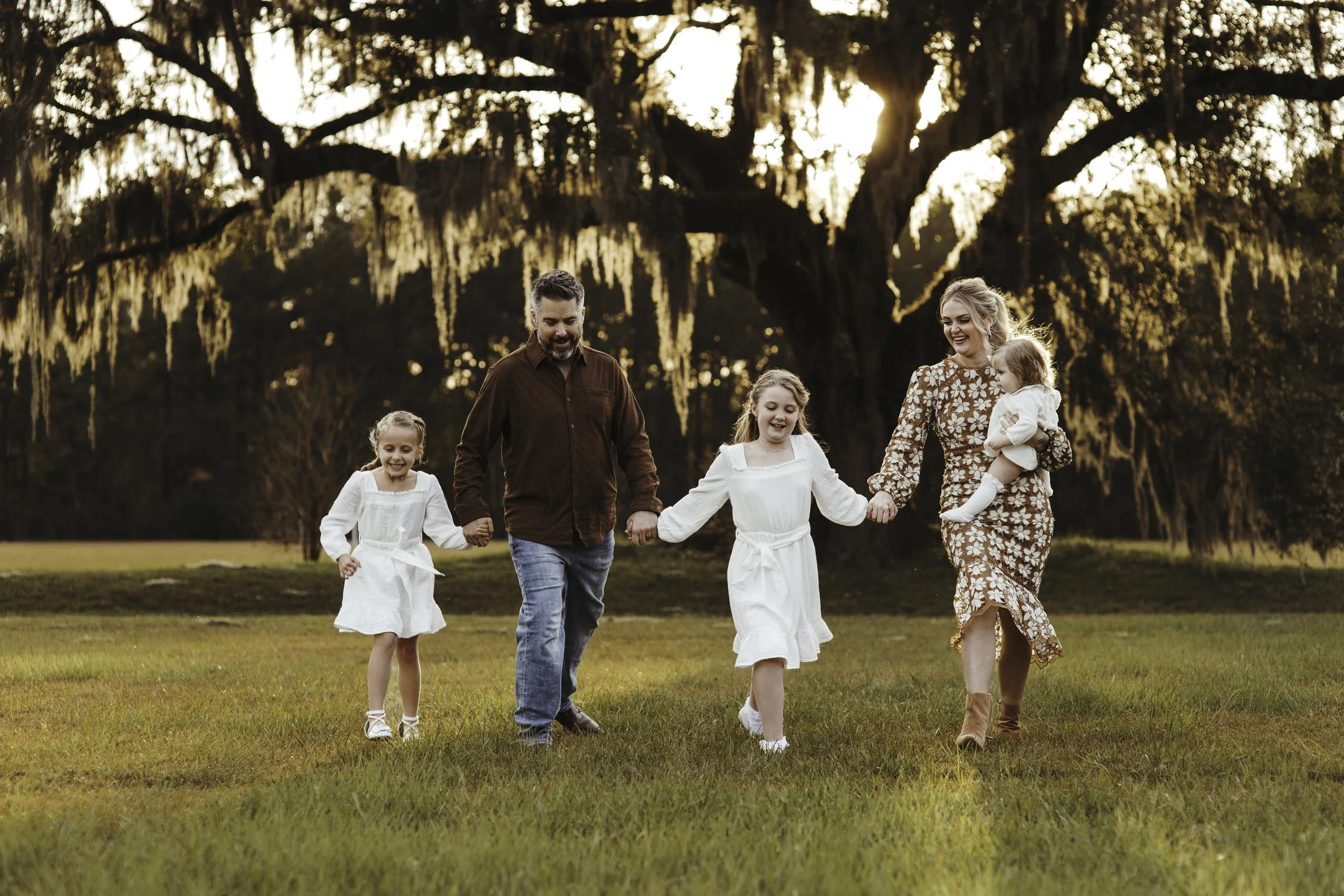 Family of five holding hands and walking on grass in a park during sunset, with large, hanging moss-covered trees in the background.