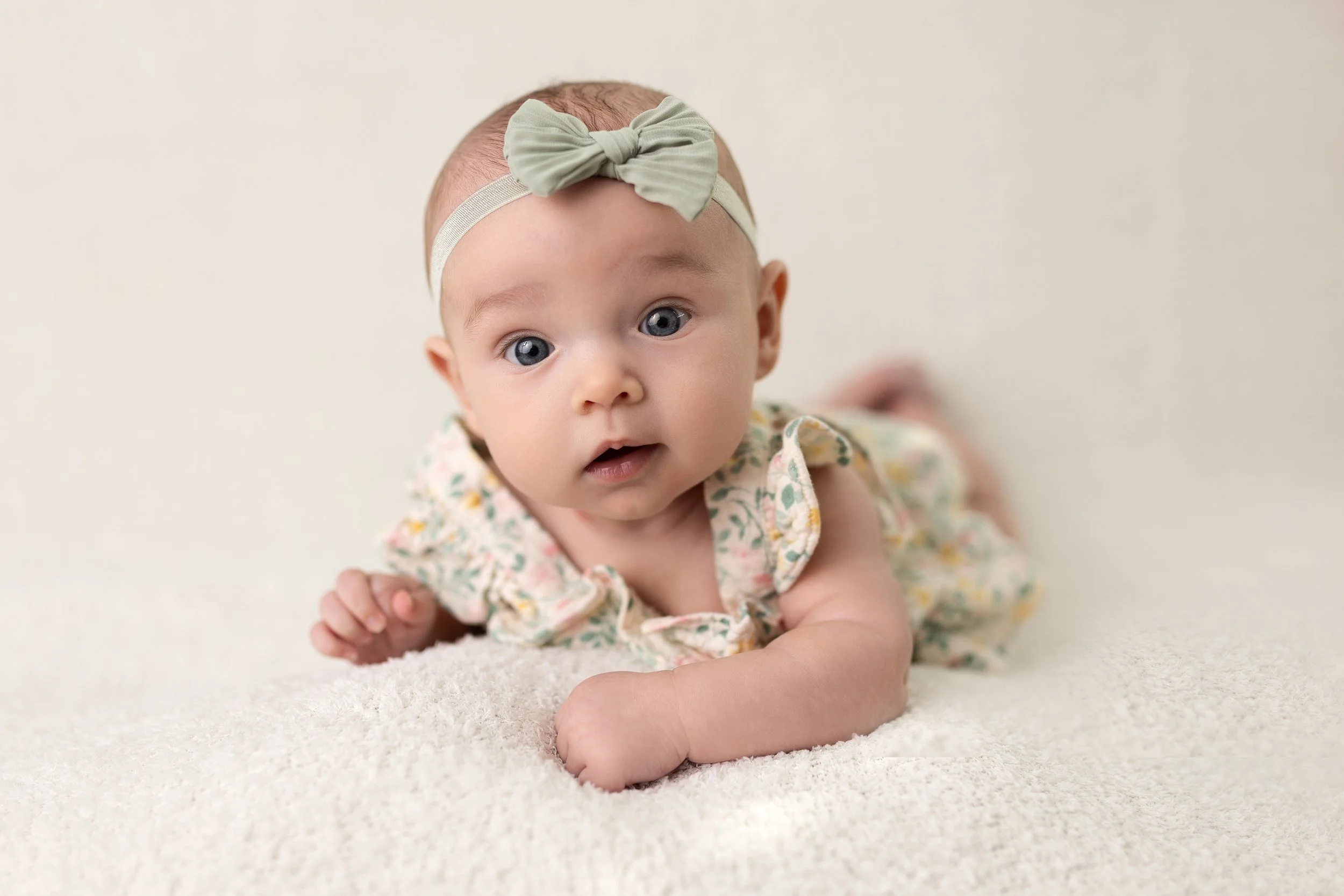 A baby girl with blue eyes, wearing a floral dress and a light green headband with a bow, lying on a soft cream-colored surface.