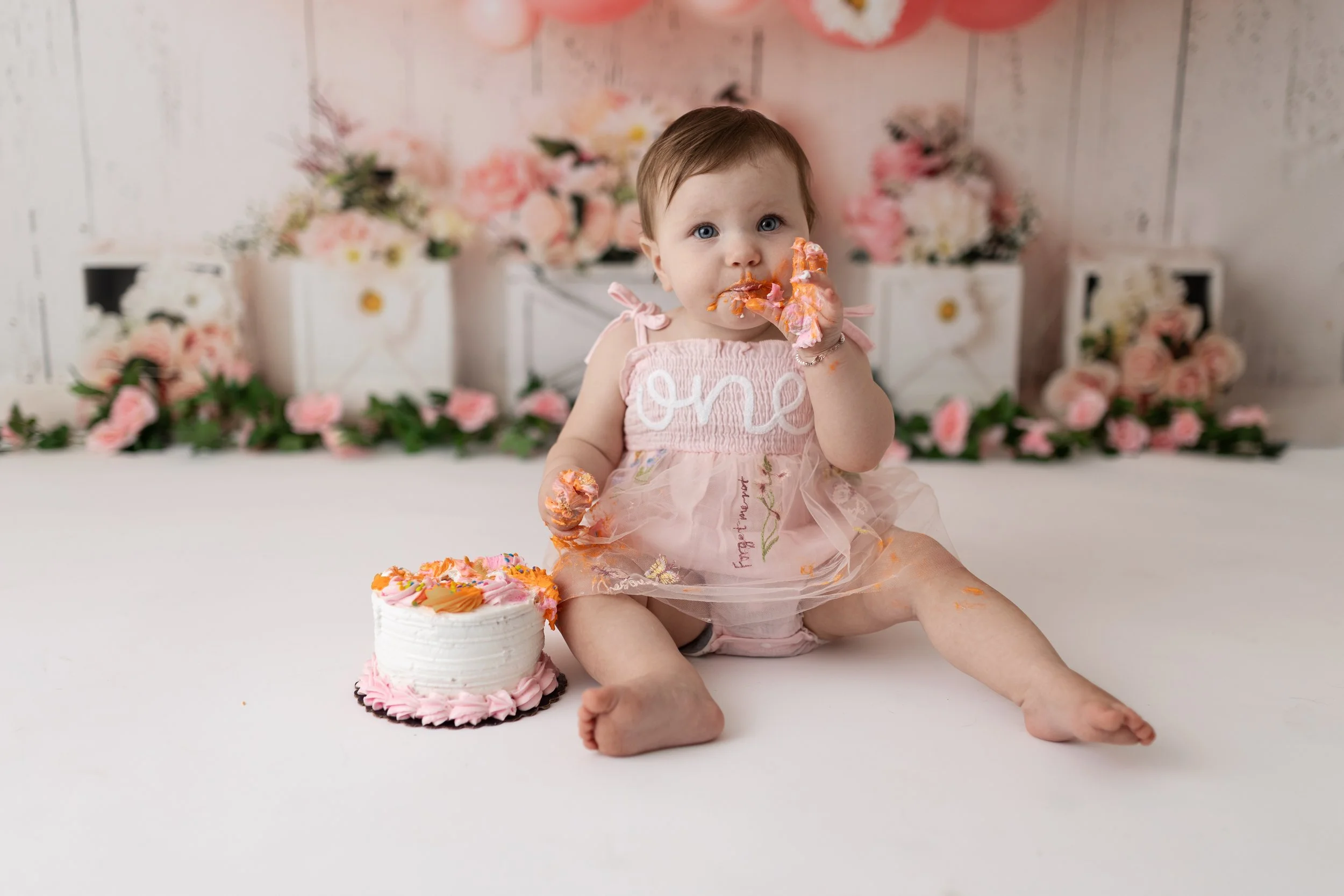 A young girl sitting on the floor with her legs spread apart, wearing a pink dress with 'one' embroidered on it, covered in pink frosting and sprinkles from a cake next to her. She is eating cake and has cake on her hands and face. The background fea