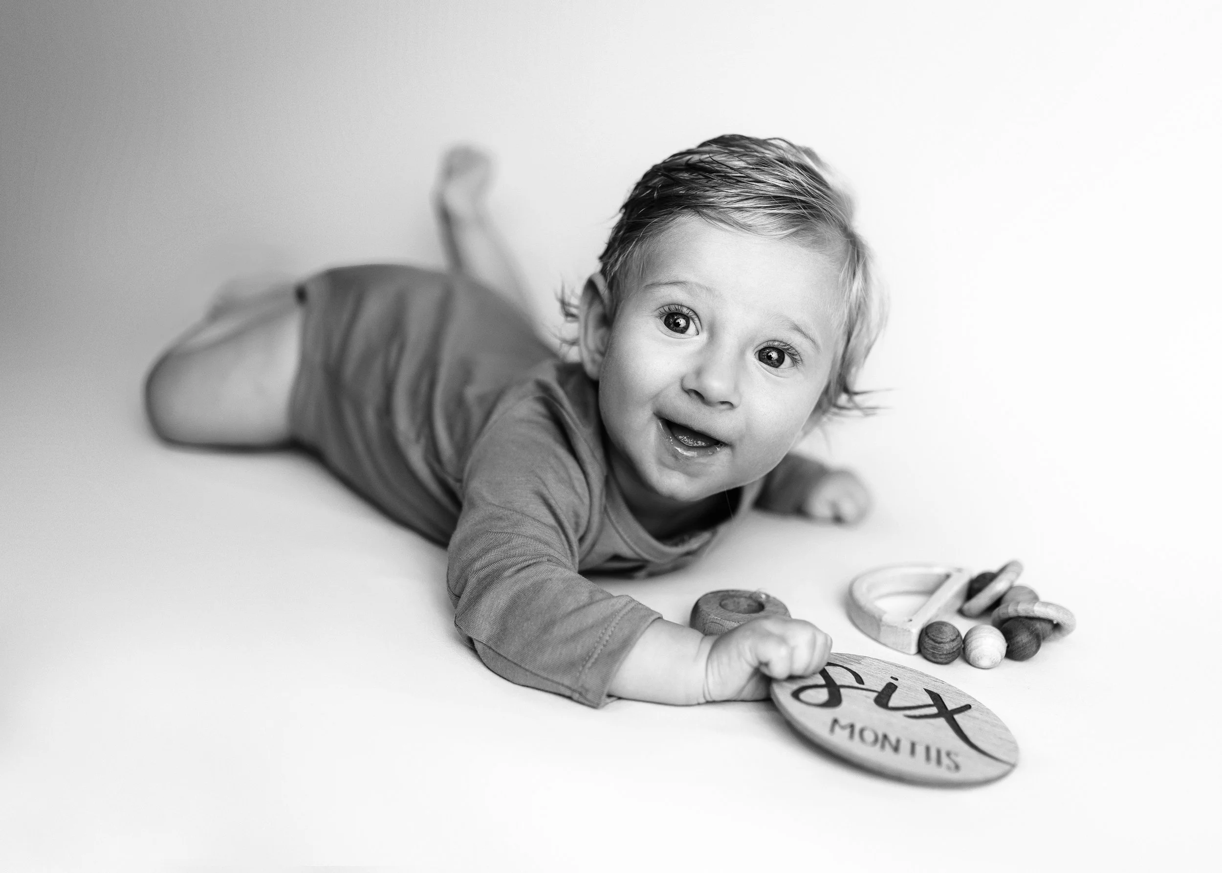 Black and white photo of a baby lying on their stomach on a plain surface, smiling and looking upward. The baby is holding a wooden sign that reads 'Six months' and has various small wooden toys scattered around.