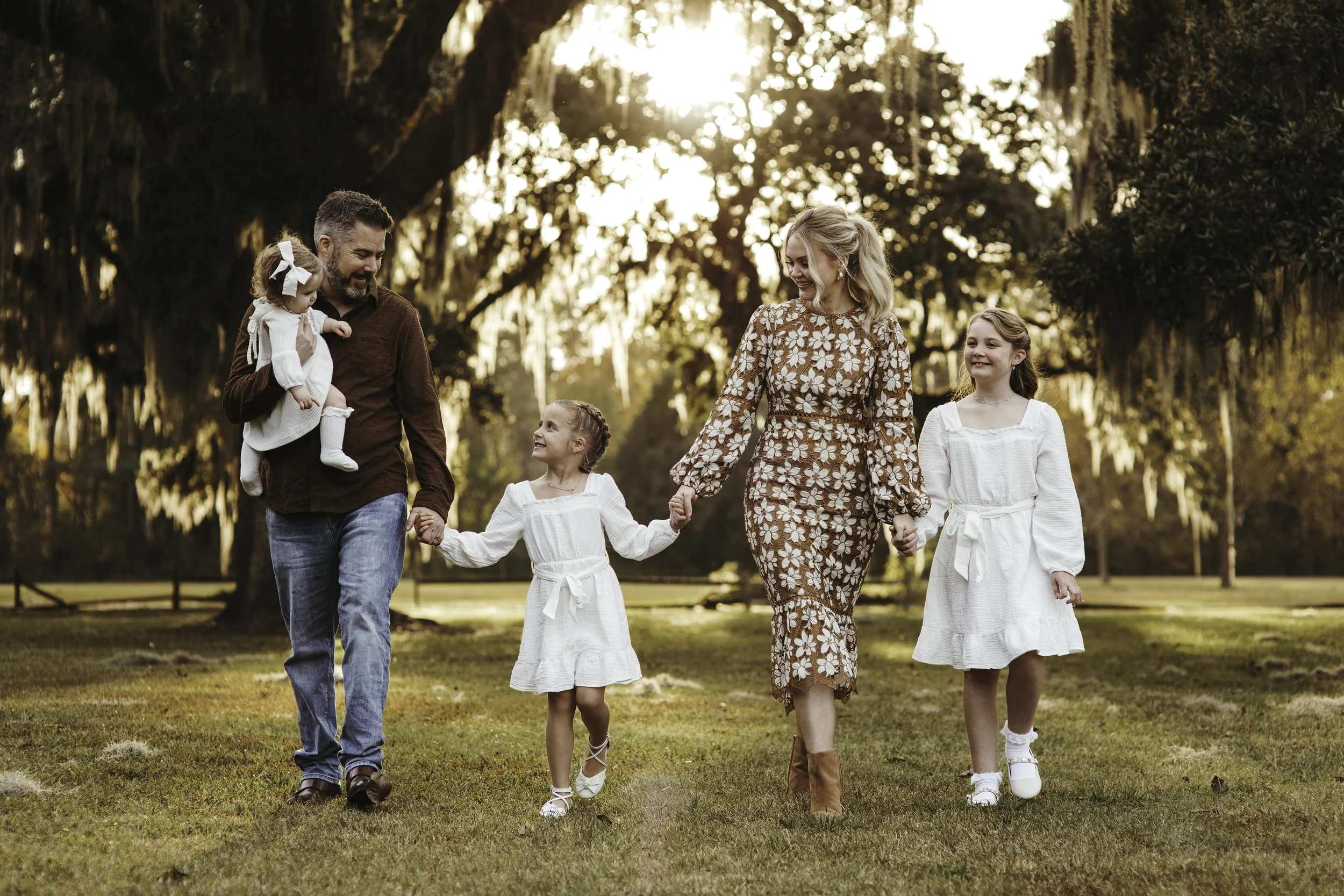 A family of five walking together outdoors in a park during sunset, holding hands and smiling. The father is carrying a young girl in white on his shoulder, the mother is holding hands with two young girls in white dresses, all four children are smil
