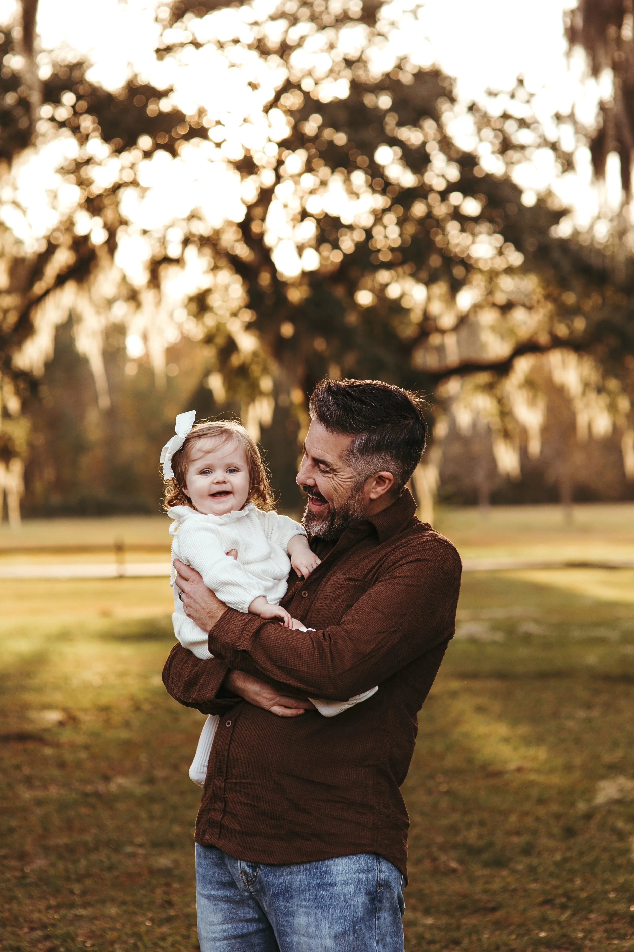A bearded man in a brown shirt holding a smiling young girl with curly hair in a white sweater outdoors during sunset, with trees and grass in the background.