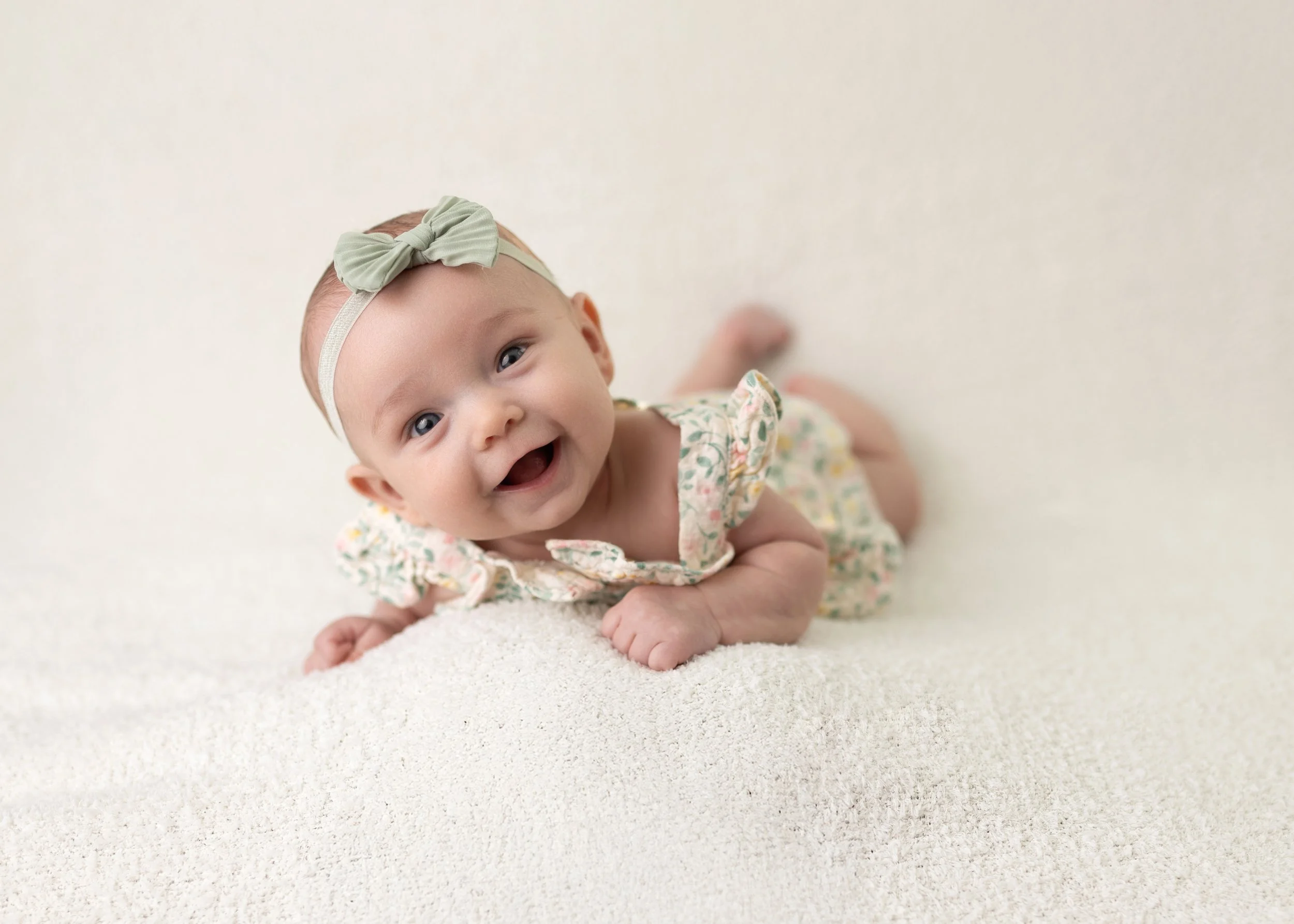 Smiling baby girl lying on her stomach on a soft, light-colored surface, wearing a floral dress and a light green headband with a bow.