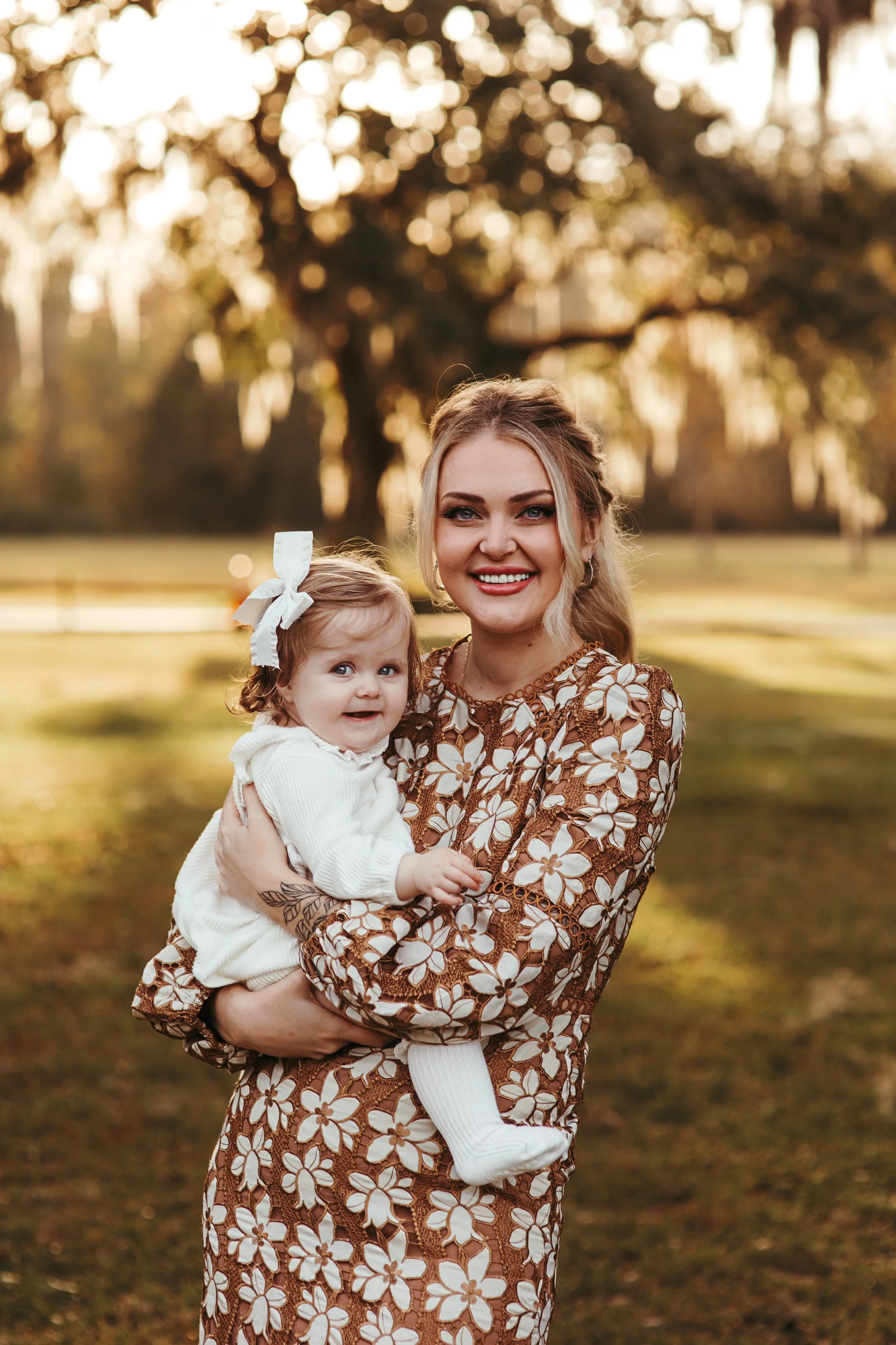 A woman holding a young girl outdoors in a park during sunset, with trees and grassy area in the background.