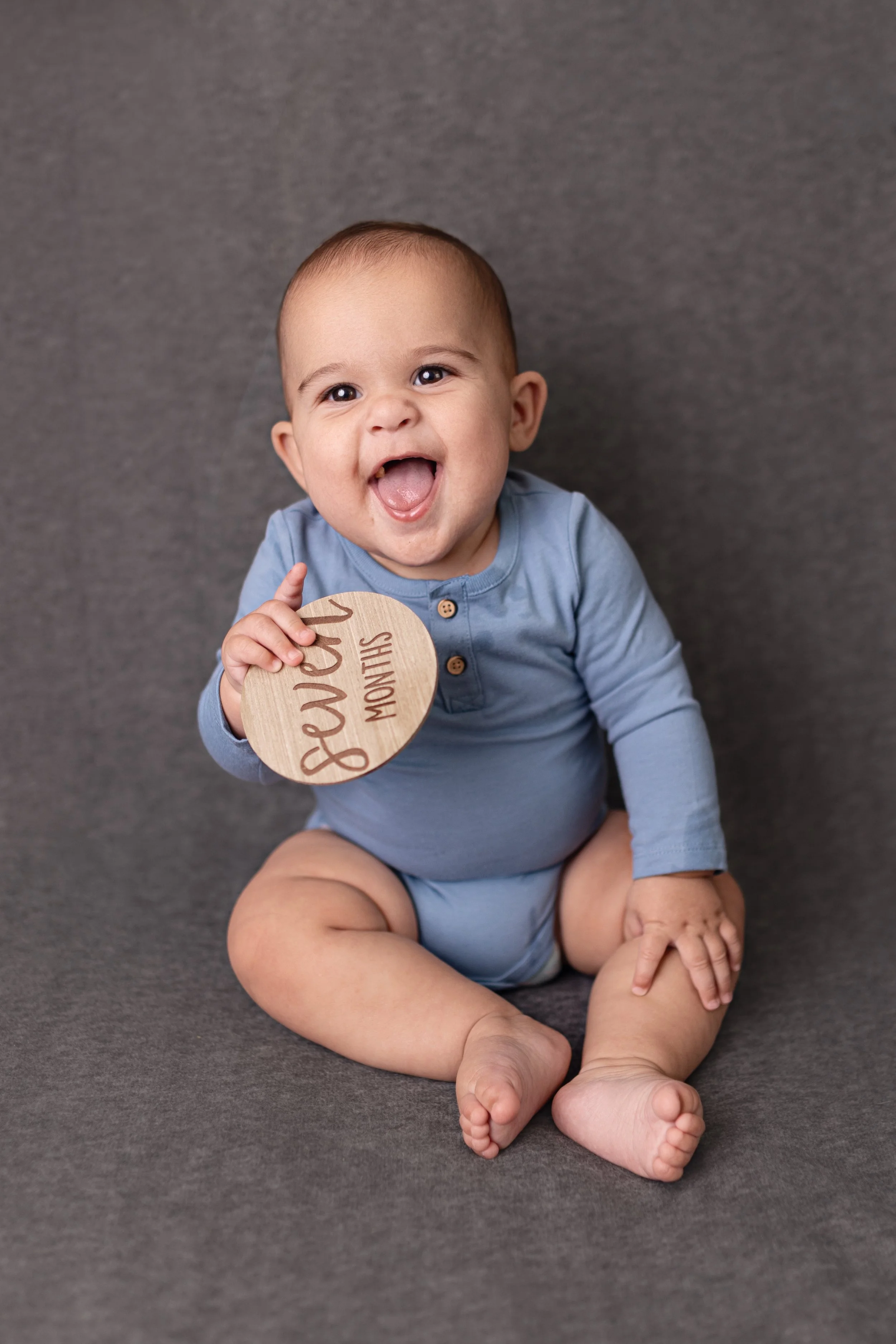 Smiling baby in blue outfit holding a wooden sign that reads 'Love 6 months' sitting on gray surface