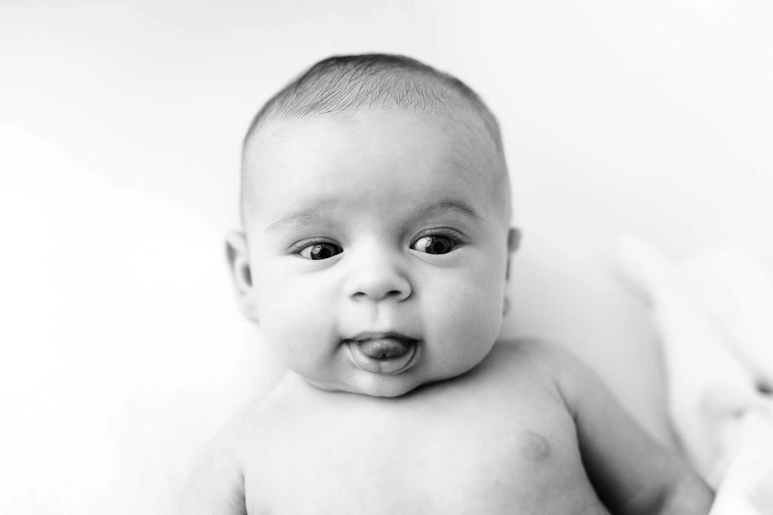 Black and white photo of a baby with short hair, wide eyes, and tongue slightly sticking out, lying on a soft surface.