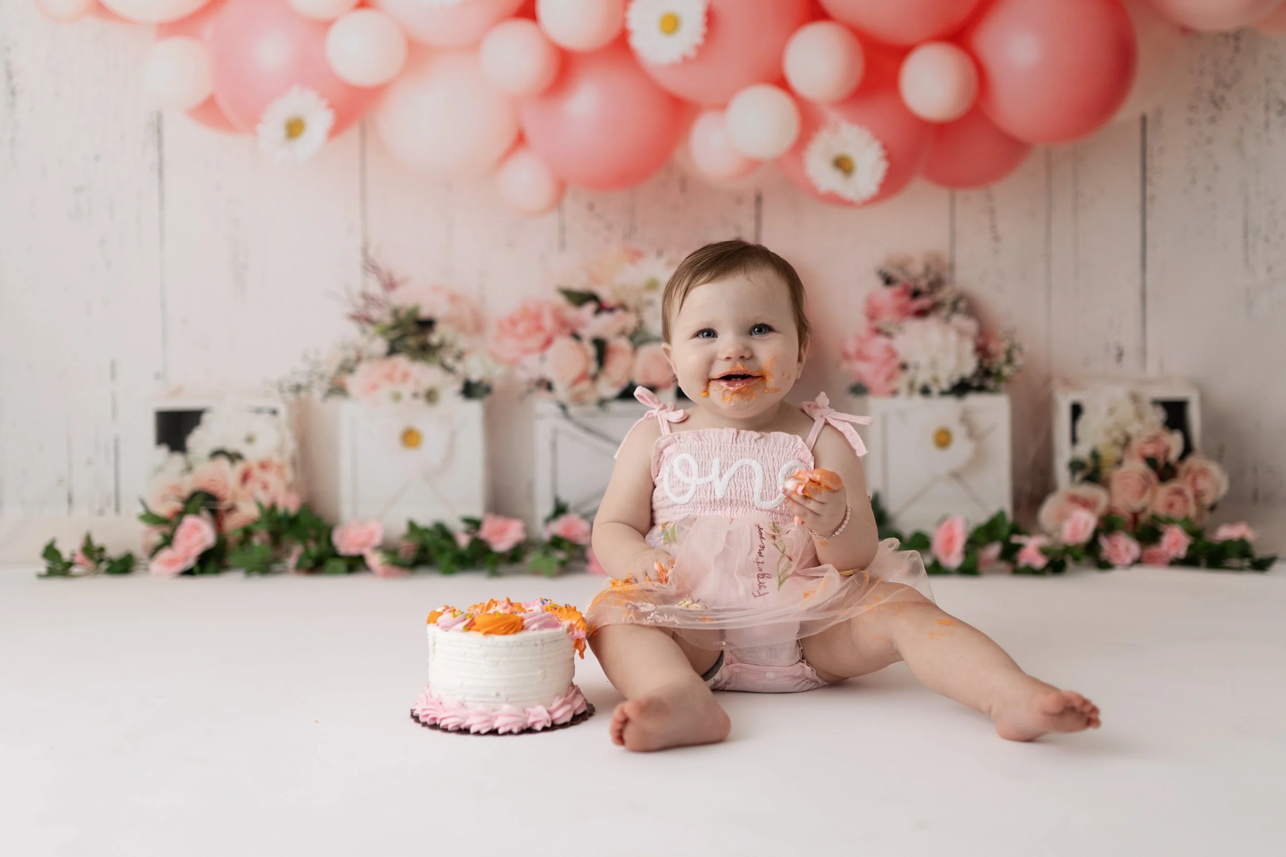 A smiling toddler girl sitting on a white floor, covered in orange frosting, celebrating her birthday with a small pink and white cake in front of her. She is wearing a light pink dress with the word "one" on it. The background features pink and whit