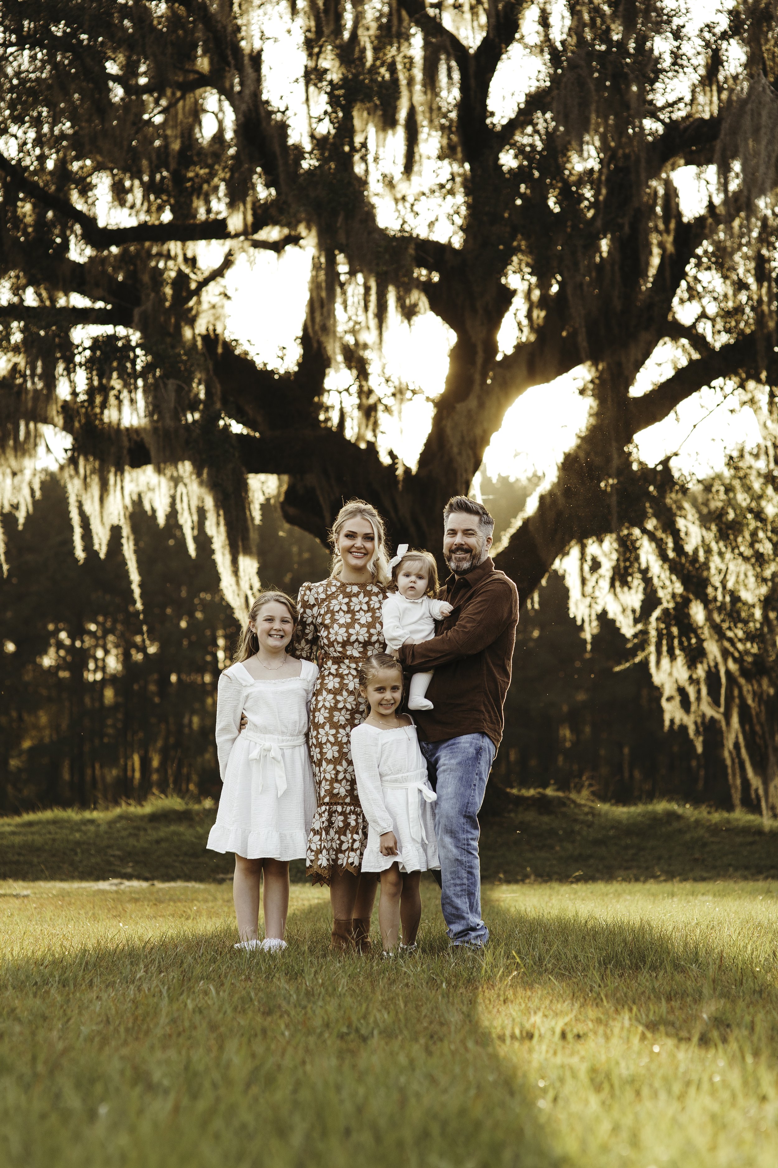 A family of five (mother, father, three daughters) standing outdoors on grass during sunset with a large tree in the background.