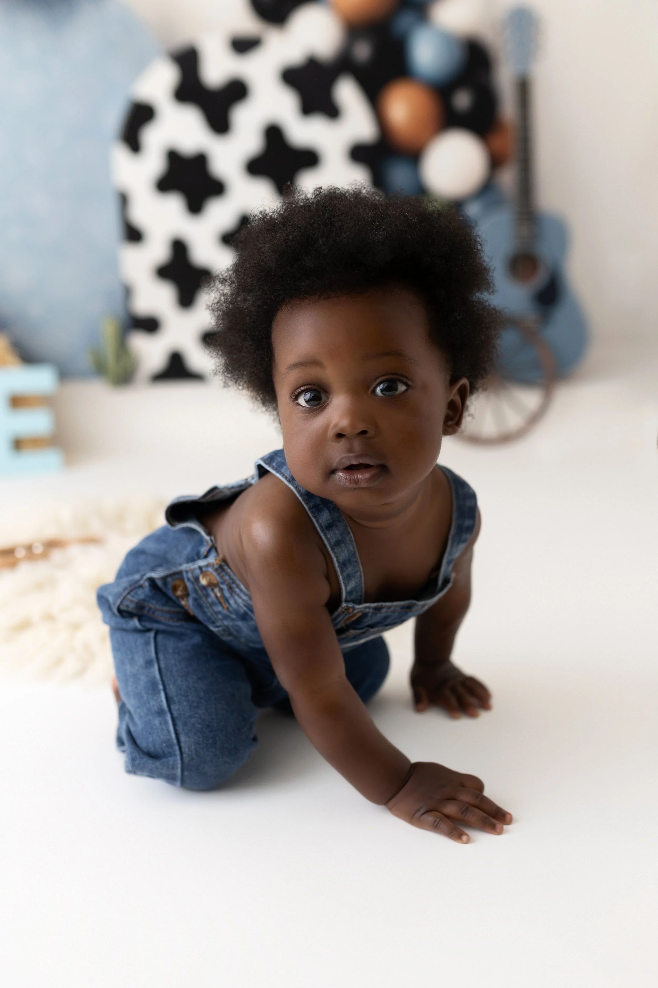 A young child with dark curly hair crawling on a white surface, wearing denim overalls, with a colorful background including a guitar, balloons, and a patterned backdrop.