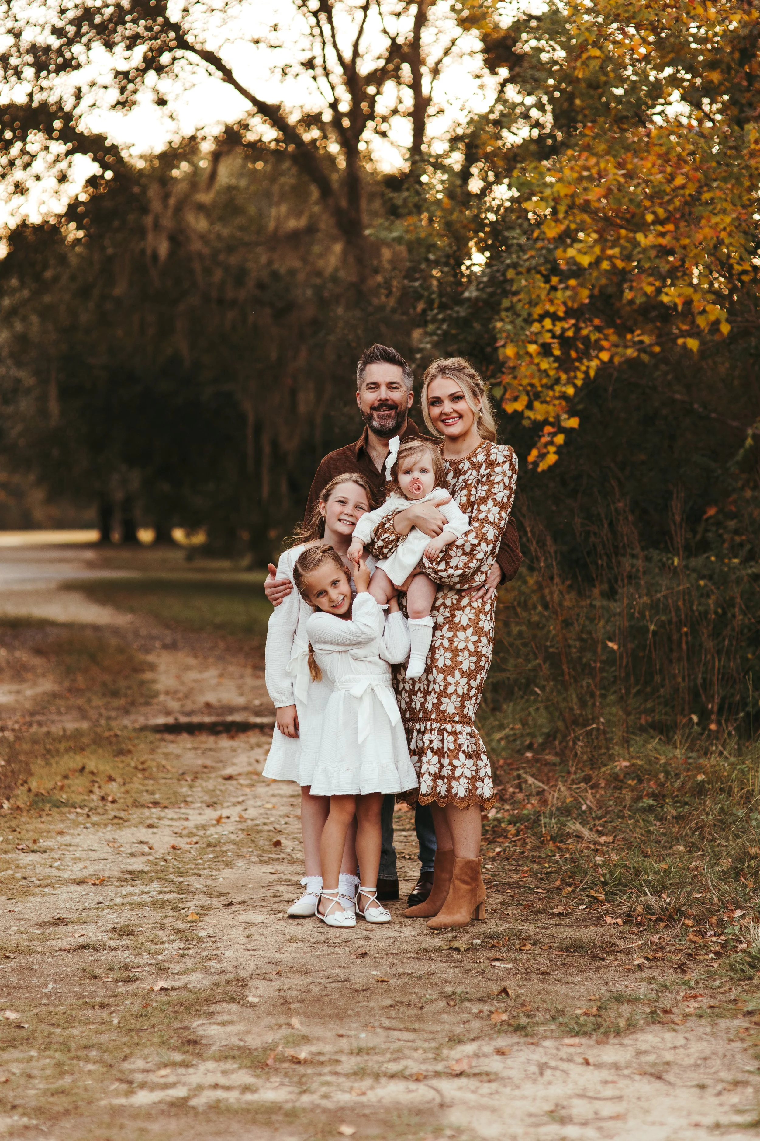 Family of five standing on a dirt path outdoors during autumn, surrounded by trees with fall foliage, smiling at the camera.