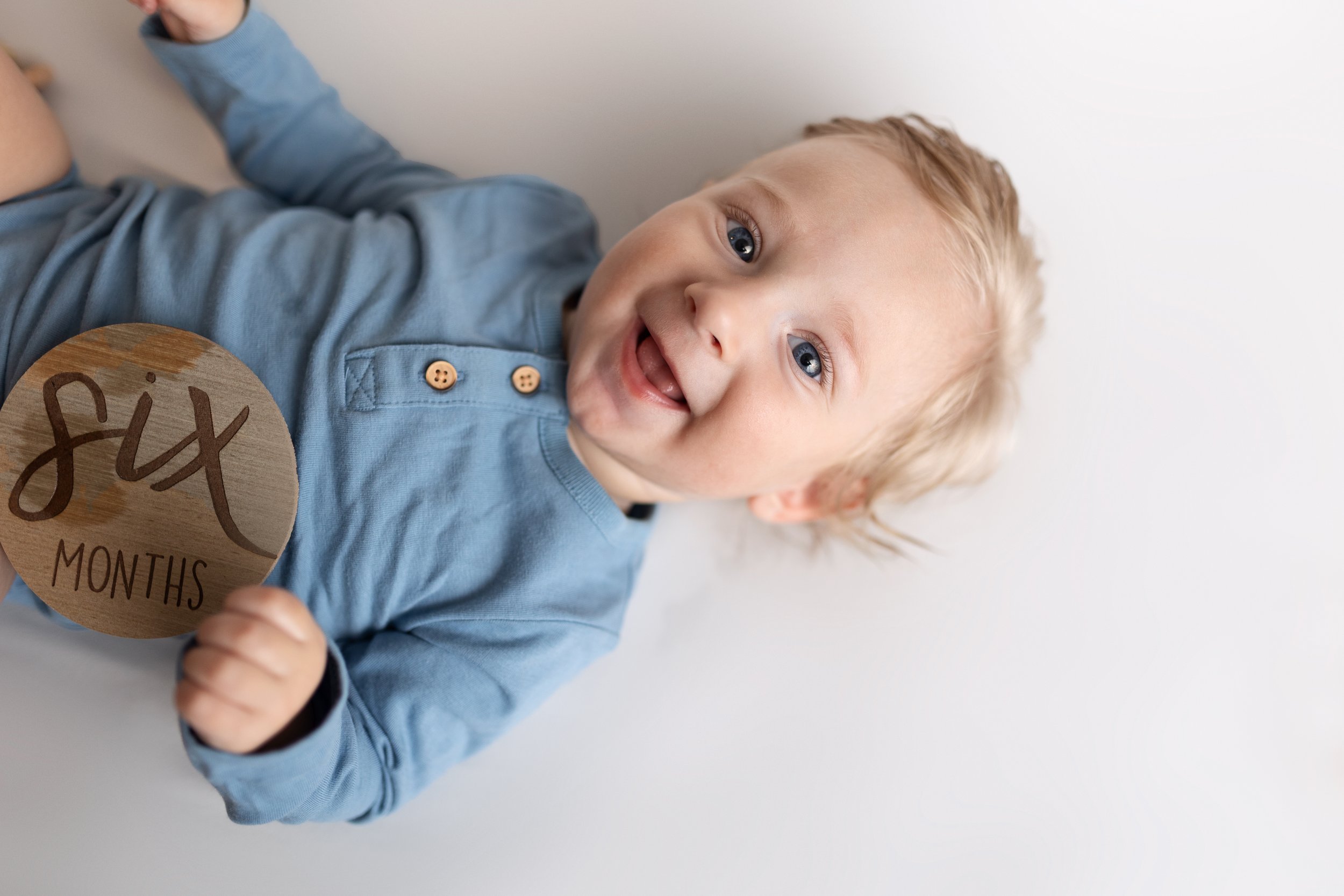 A smiling young child with blonde hair and blue eyes holding a wooden sign that reads 'six months'.