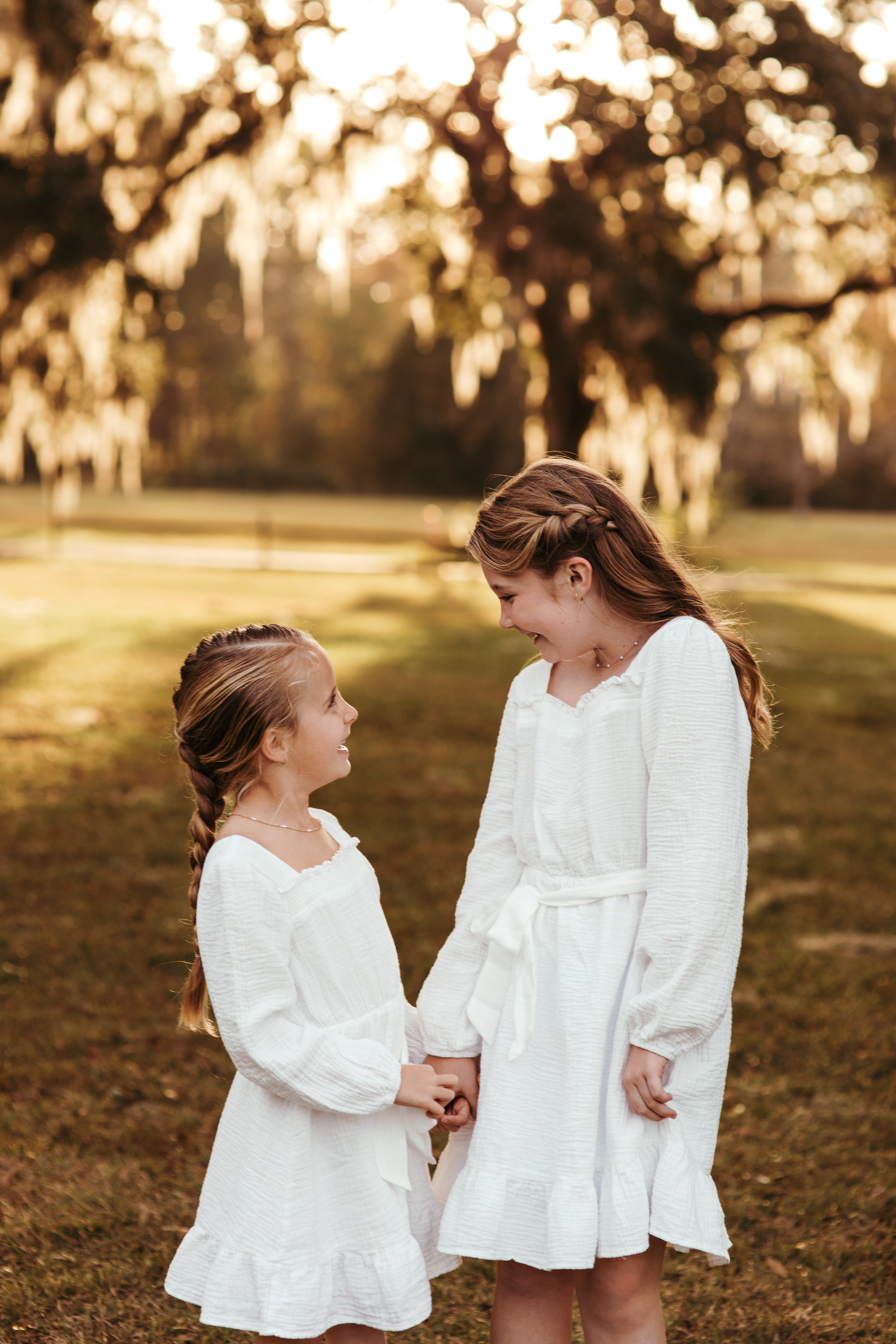Two young girls in white dresses holding hands and smiling in a park during sunset.