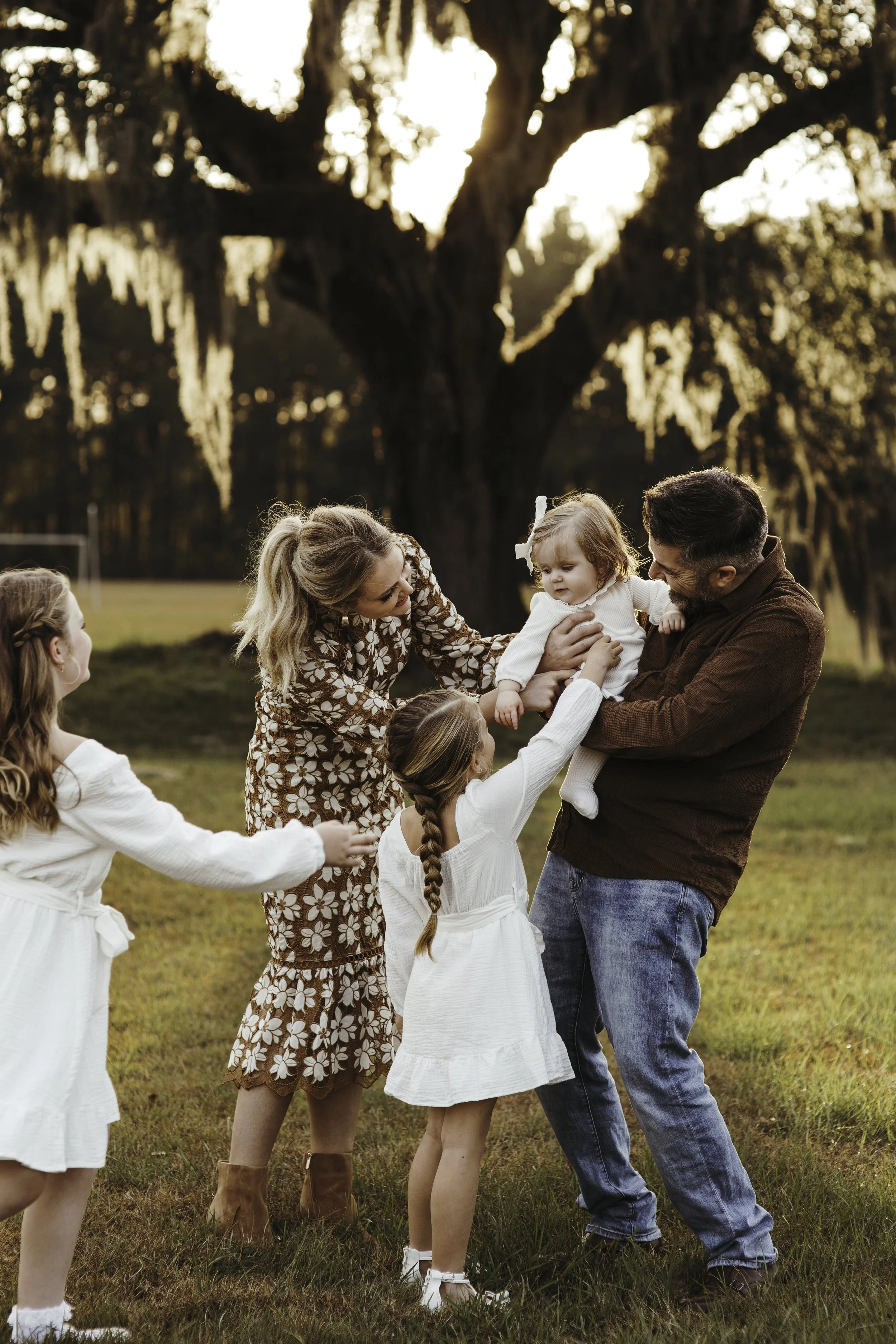 A family of five, with two adult women, one man, and three young girls, playing together outdoors in a grassy field during sunset, with a large tree in the background.