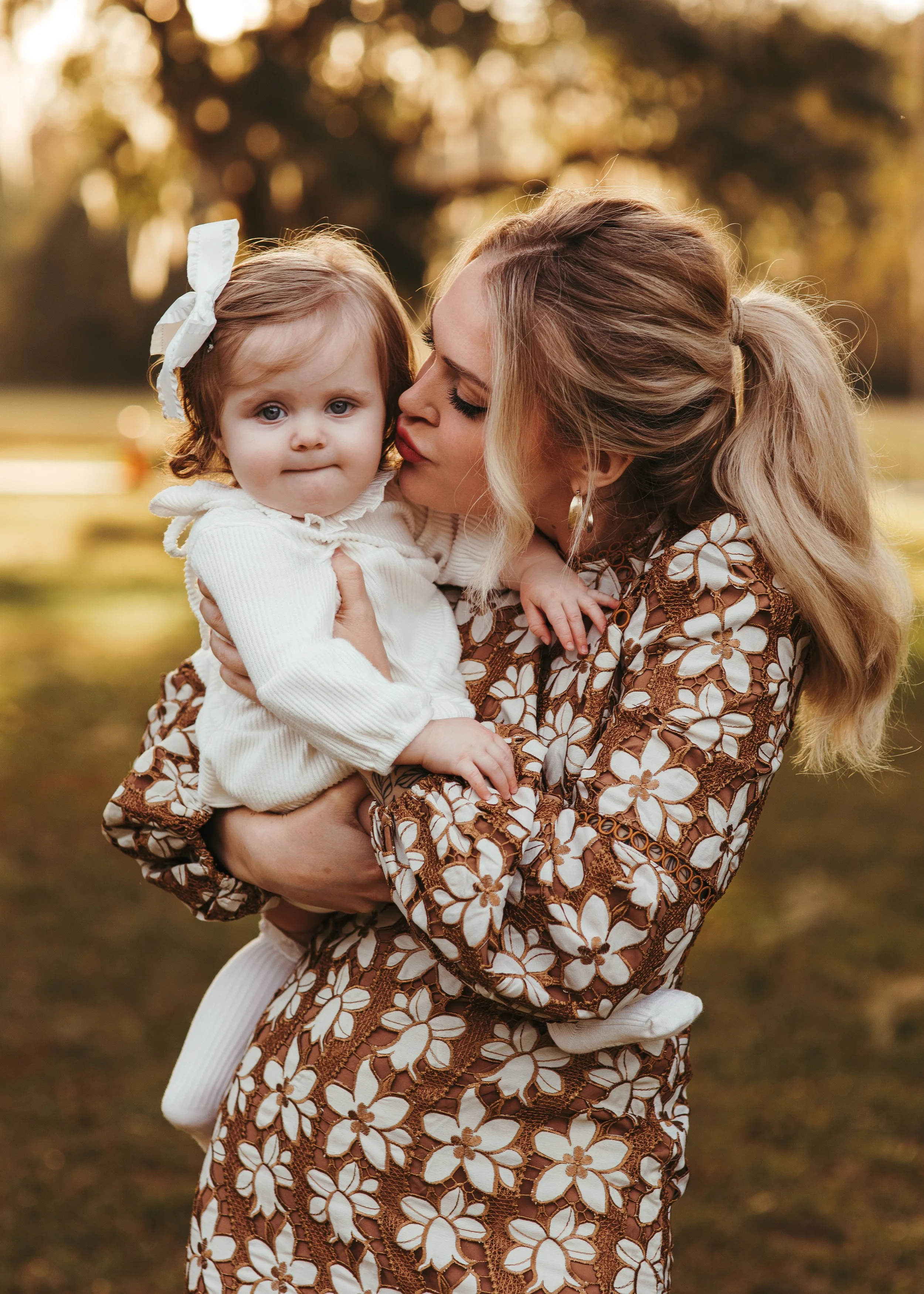 A woman holding a young girl outdoors during fall, with trees and a grassy area in the background.
