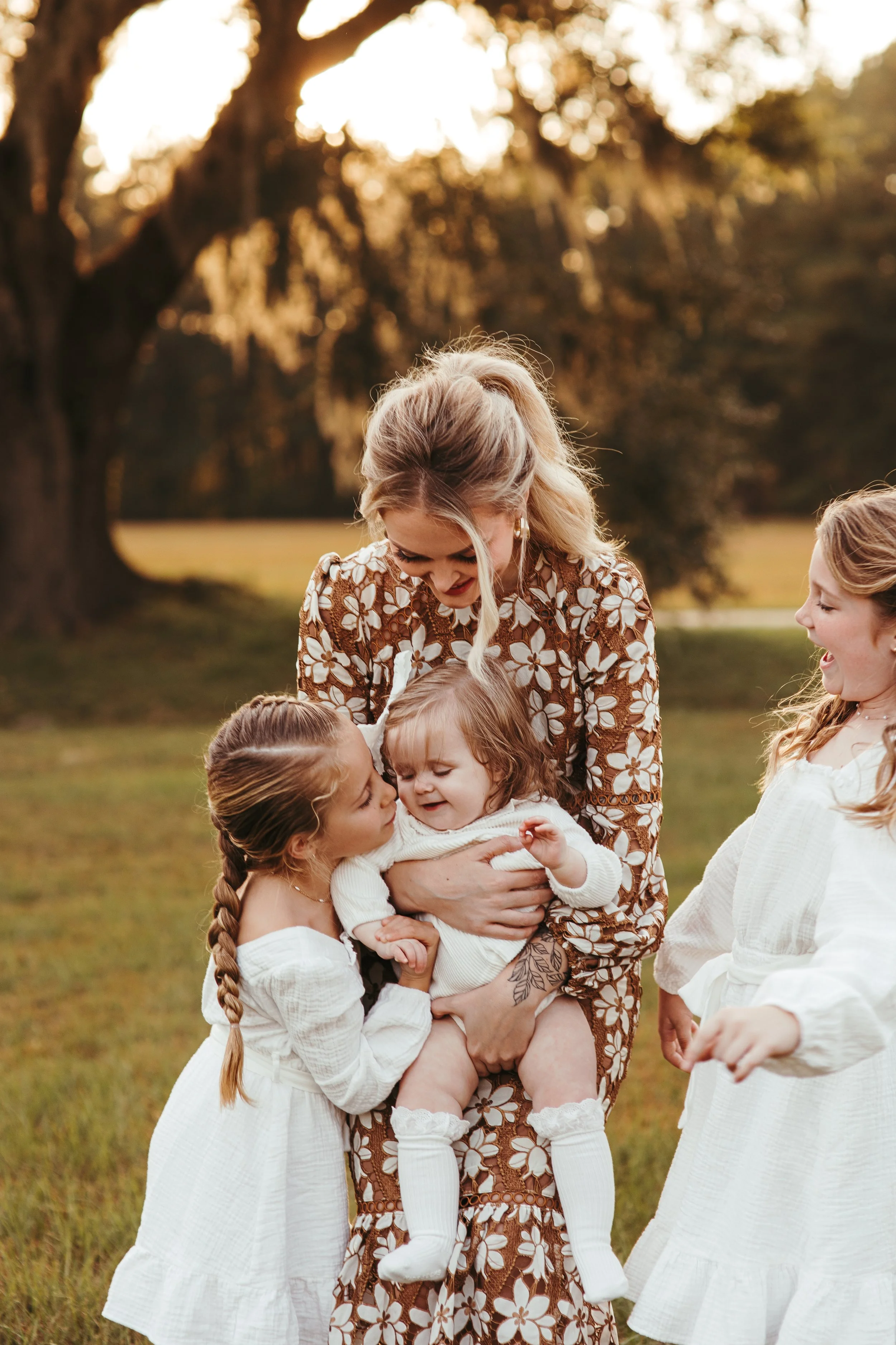 A woman holding a toddler on her lap, surrounded by two young girls, outdoors during sunset with trees and grass in the background.