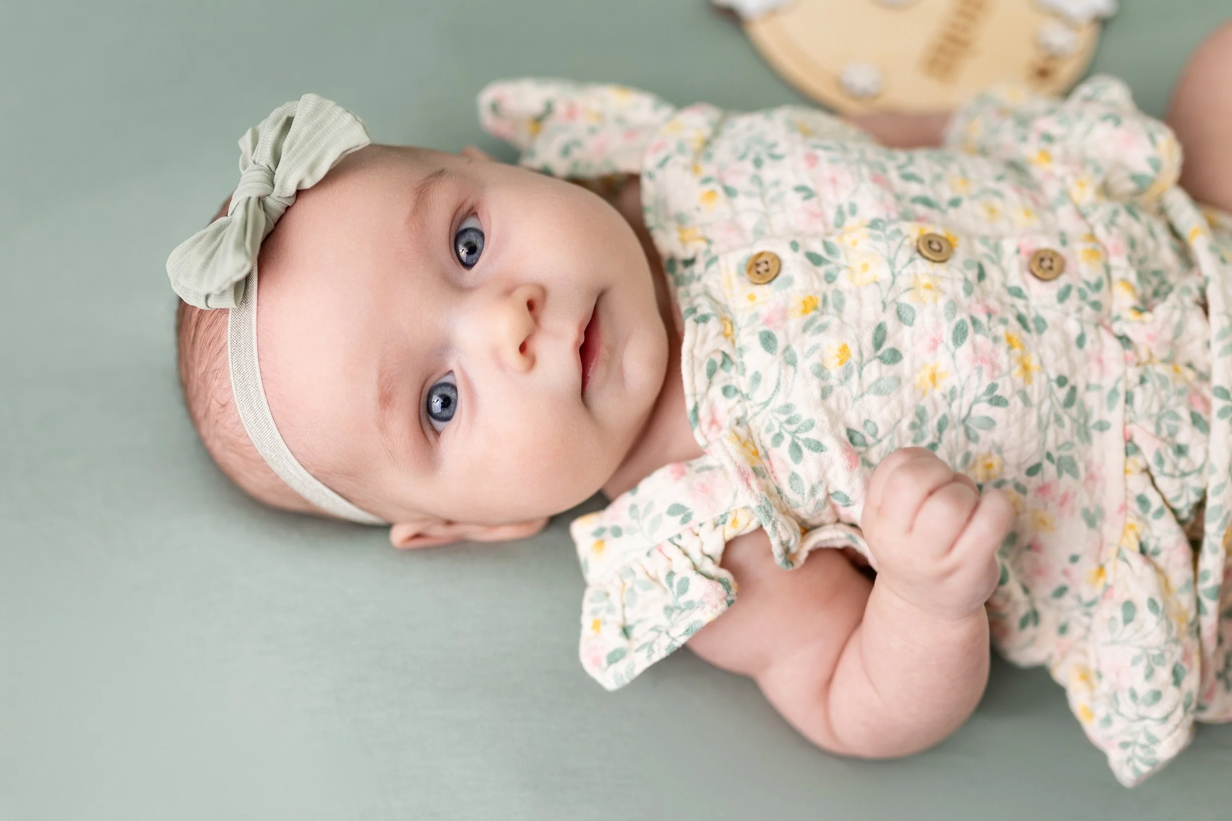 A baby girl with blue eyes lying on a green surface, wearing a floral dress and a light green bow headband.